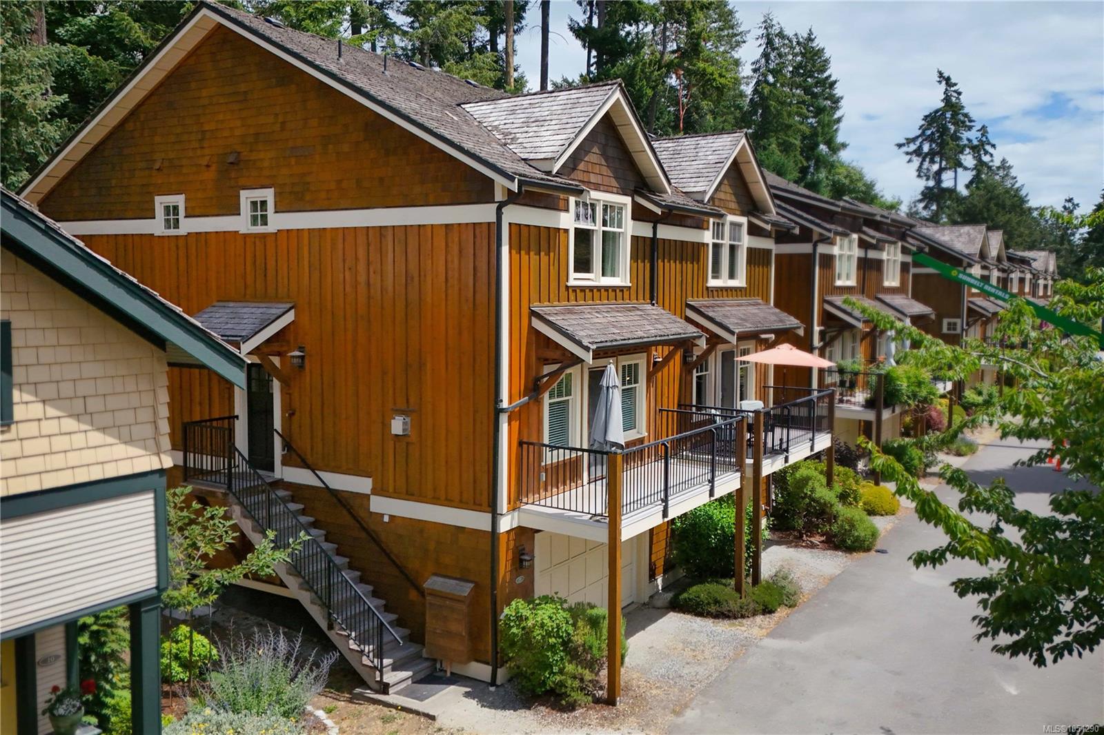 Attached wood-sided homes with gabled roofs, raised balconies, and exterior stairs beside a leafy lane.