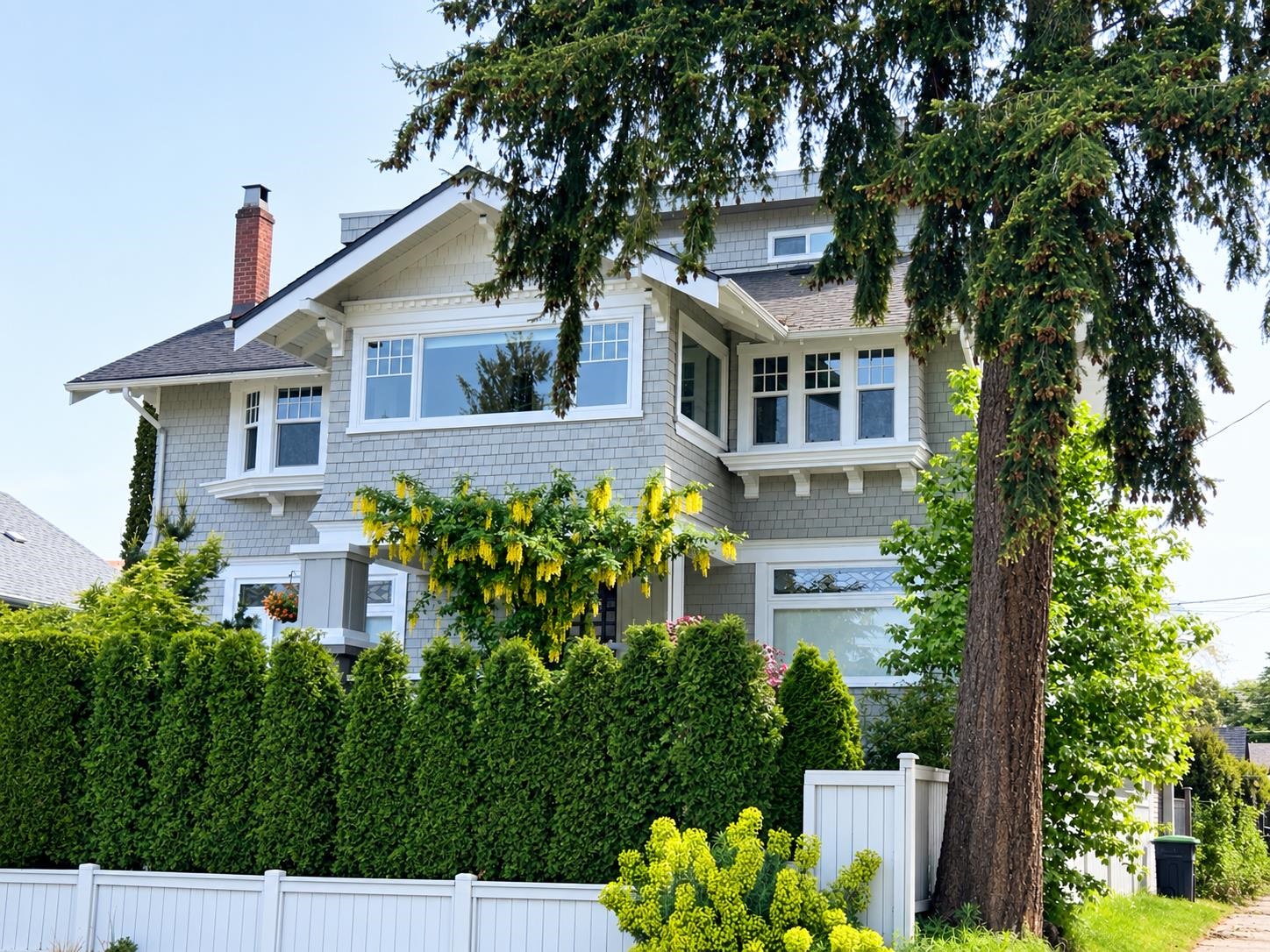 Gray shingle-sided house with white trim and brick chimney, framed by mature trees, hedges, and a white fence.