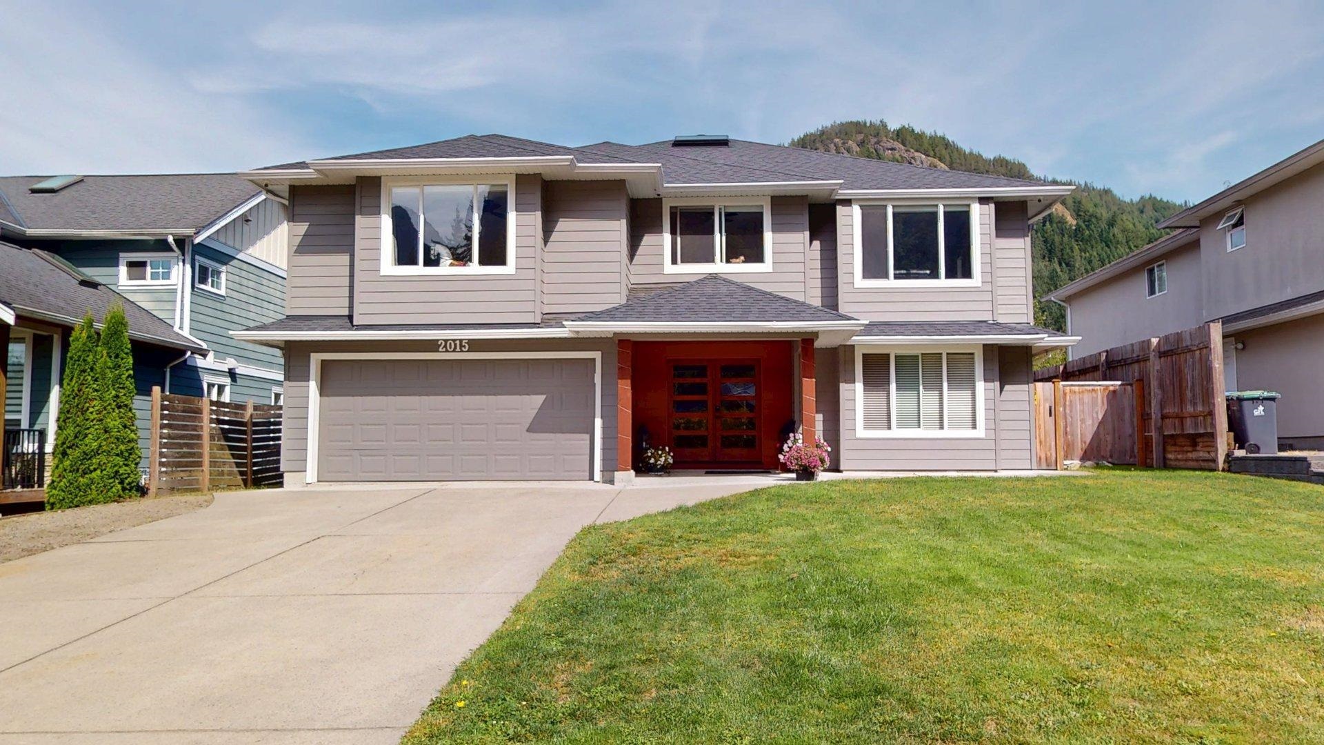 Two-story gray home with a red entrance, double garage, and forested hills in the background.