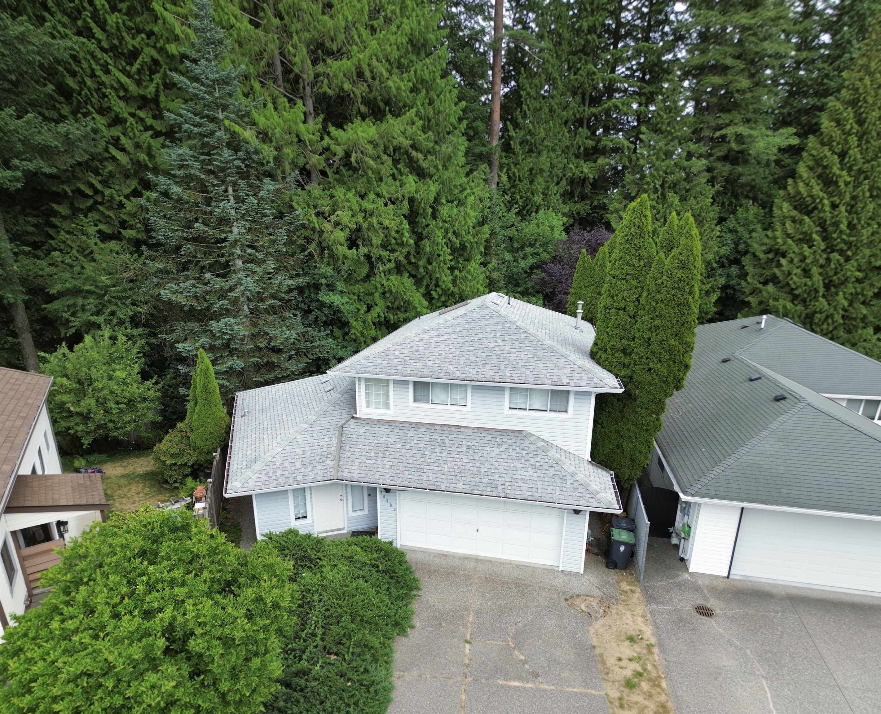 Aerial view of a white two-story house with a gray roof, front garage, driveway, and surrounding evergreen trees.