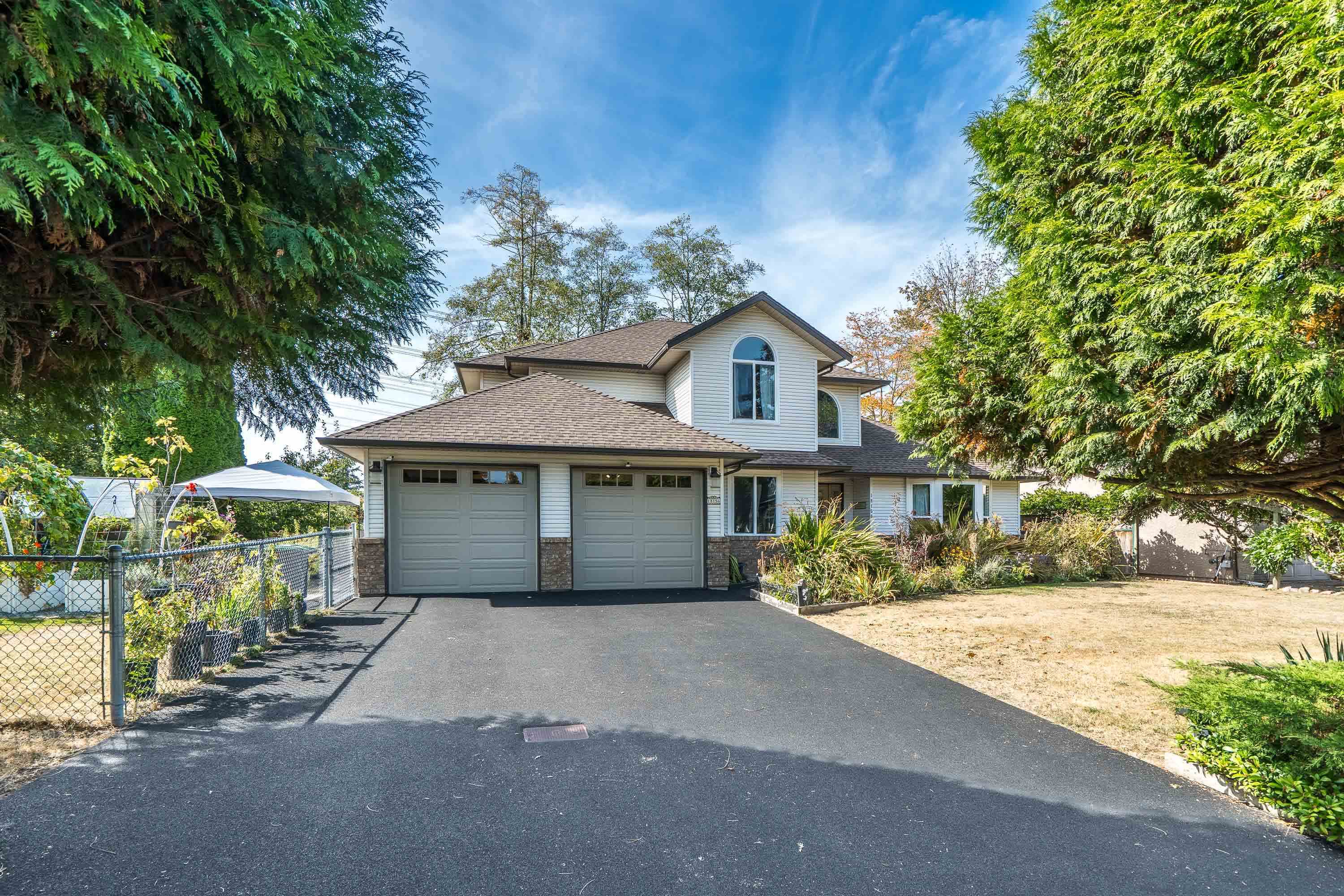 Two-story white house with a double garage, wide driveway, garden beds, and mature evergreen trees.