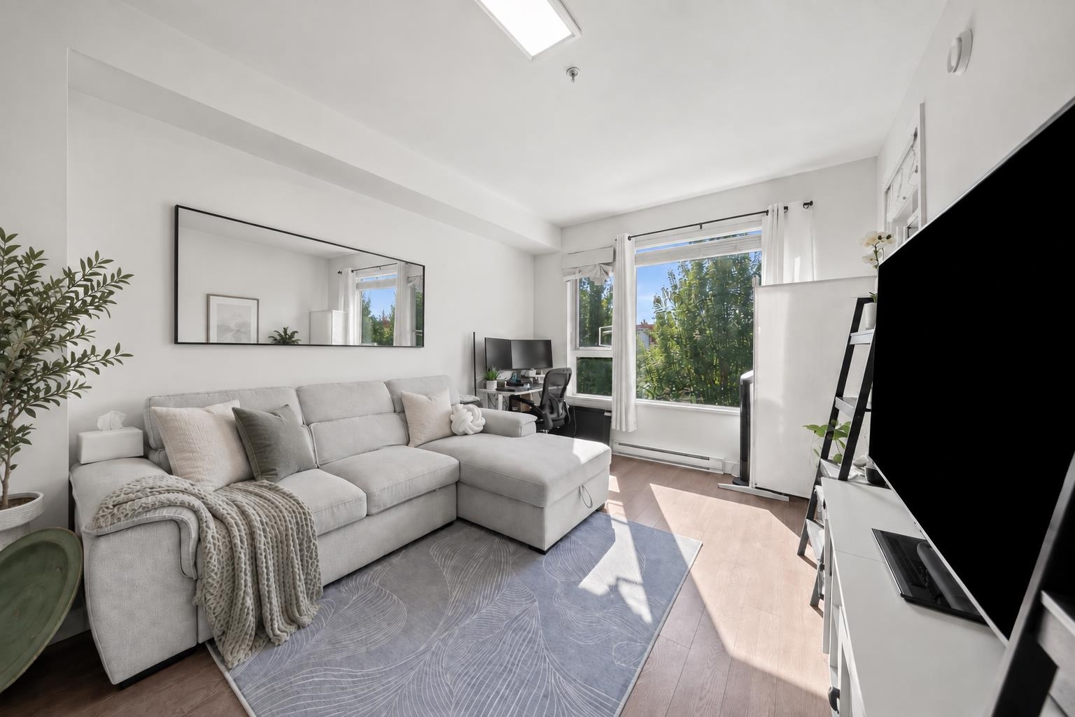 Bright living room with a gray sectional sofa, patterned rug, and tall windows overlooking greenery.
