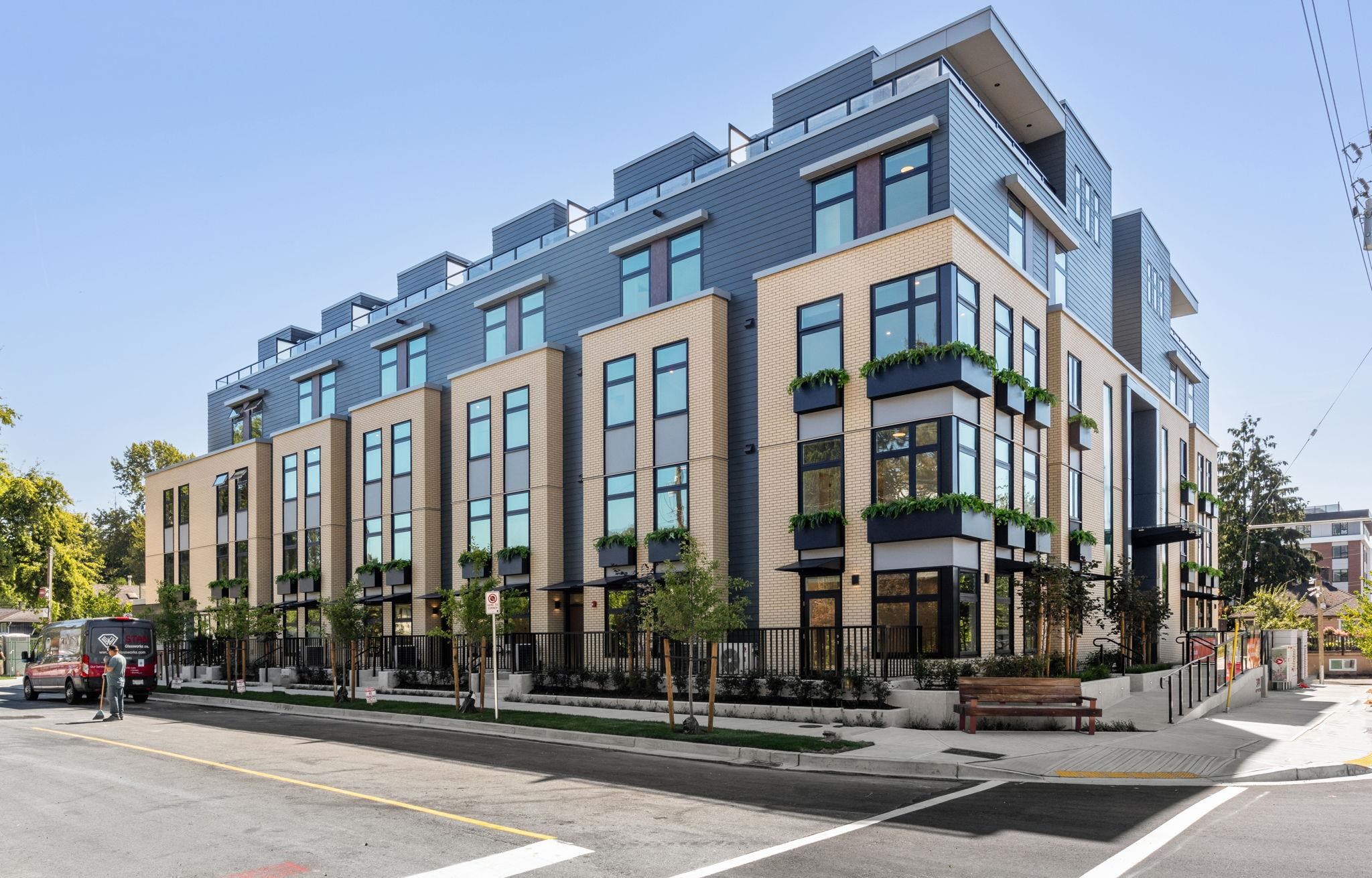 Modern multi-unit residential building with tan brick, blue-gray siding, and planted balconies.