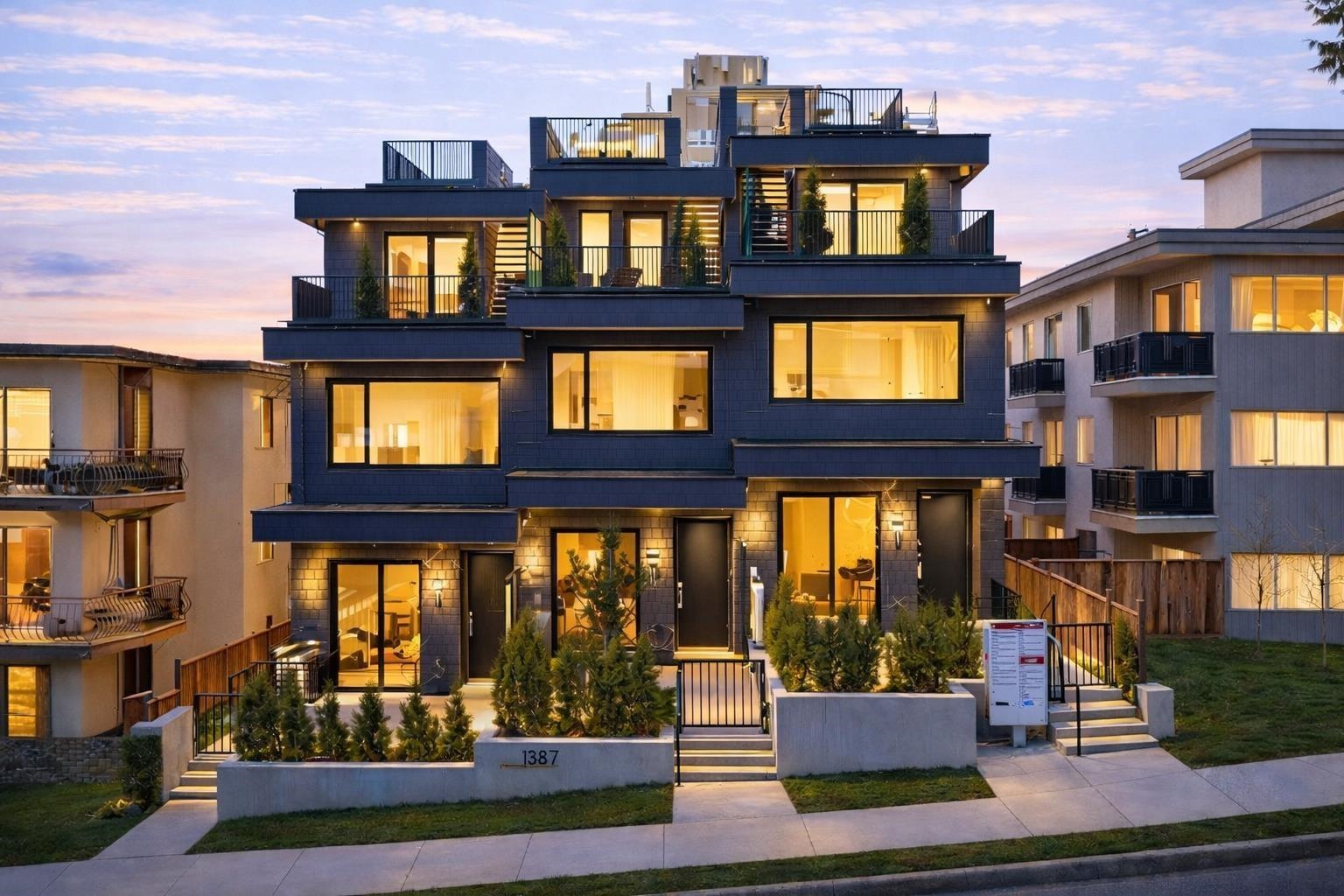 Modern multi-story residence with rooftop terraces, illuminated windows, and landscaped entrance at dusk.