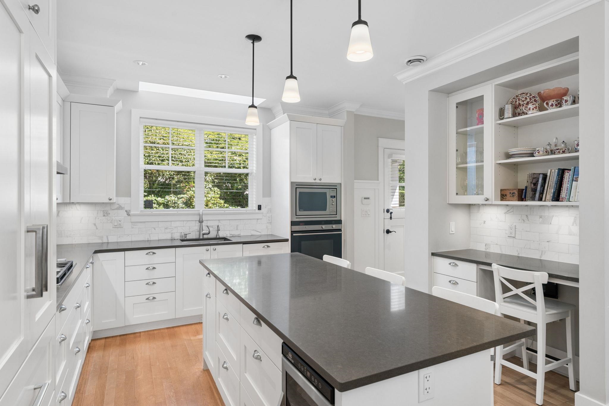 Bright white kitchen with a large dark island, marble-look backsplash, and window overlooking greenery.