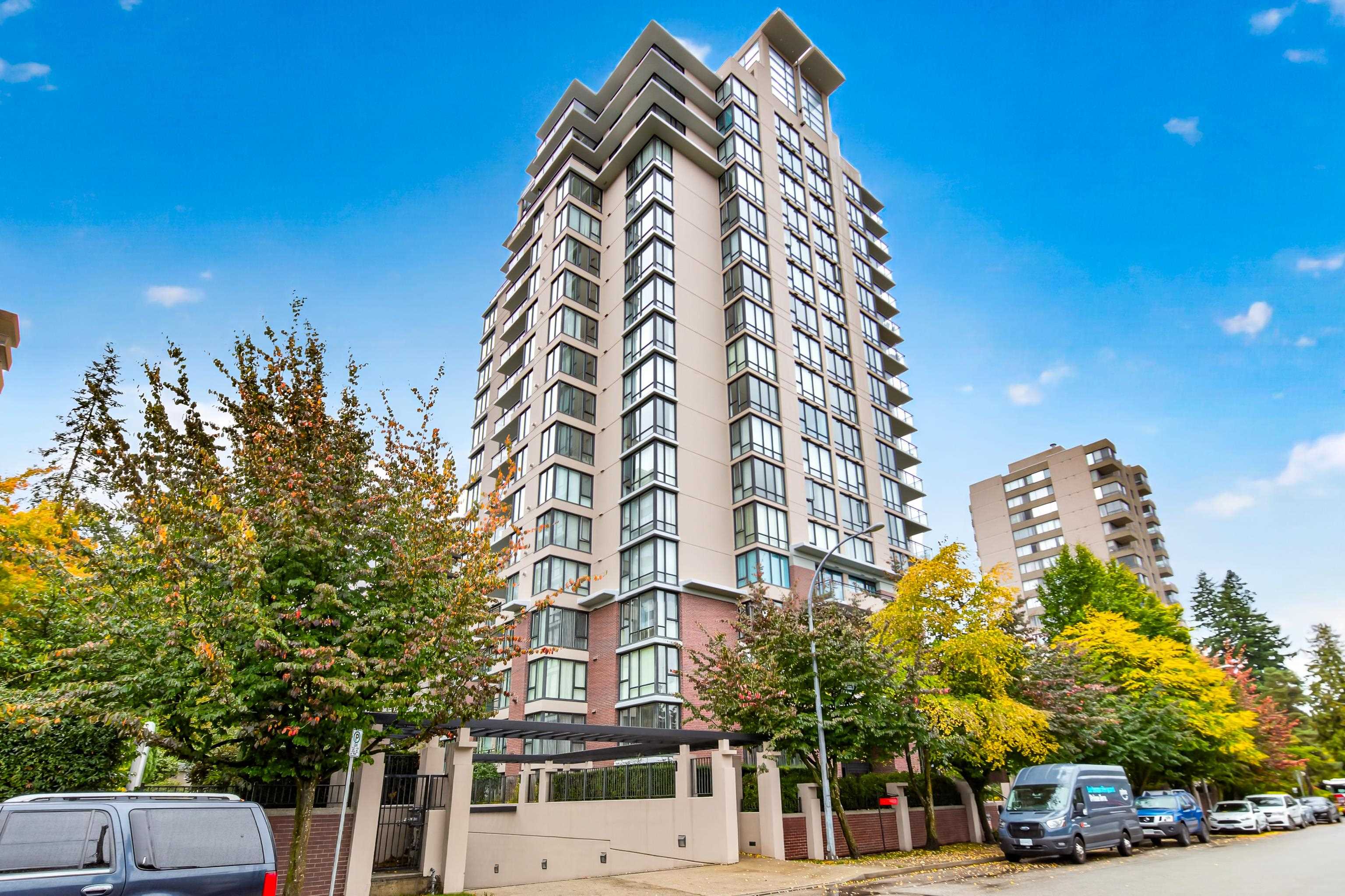Modern condominium tower with balconies, framed by autumn trees along a street.