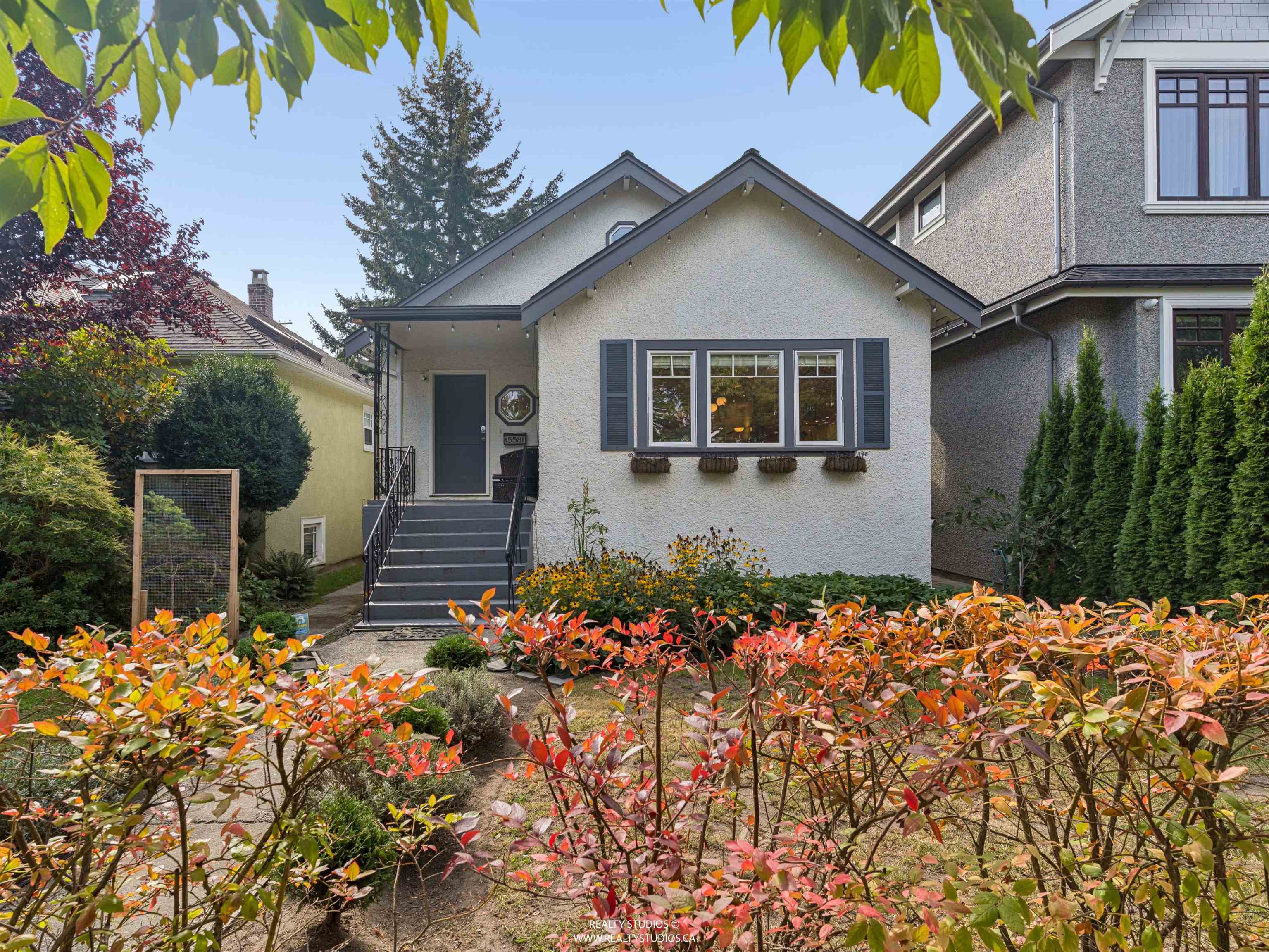 Small light-colored house with broad front steps, gray shutters, and red-orange autumn foliage in front.