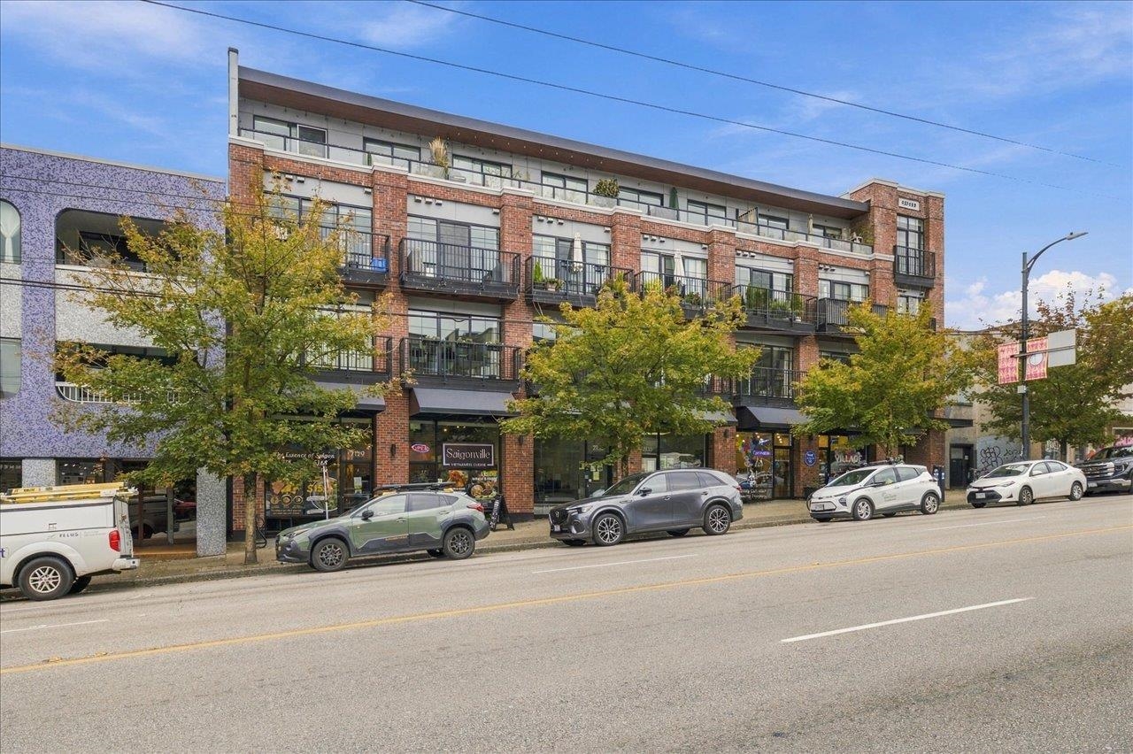 Mixed-use brick building with balconies above street-level shops and parked cars along the curb.