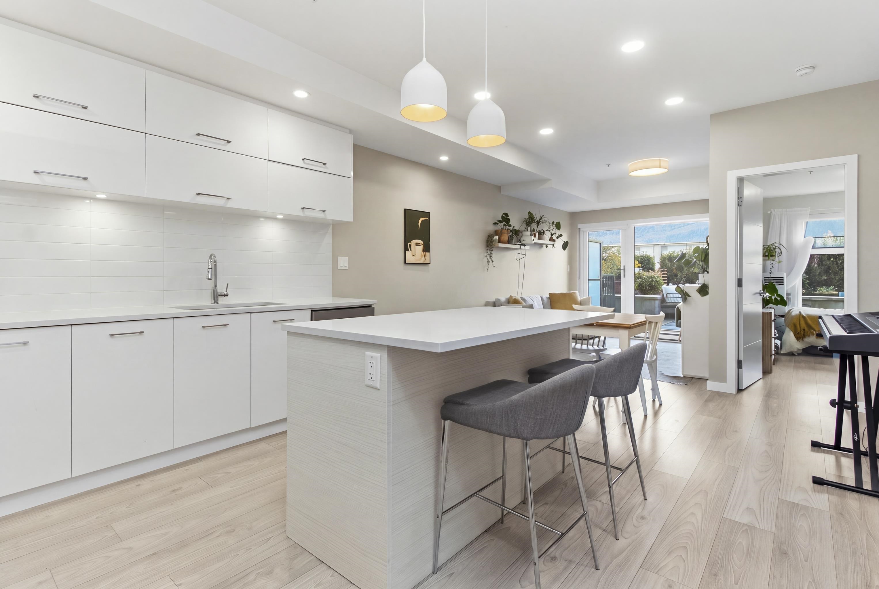 Modern white kitchen with a large island and bar stools, opening onto a living area and balcony.