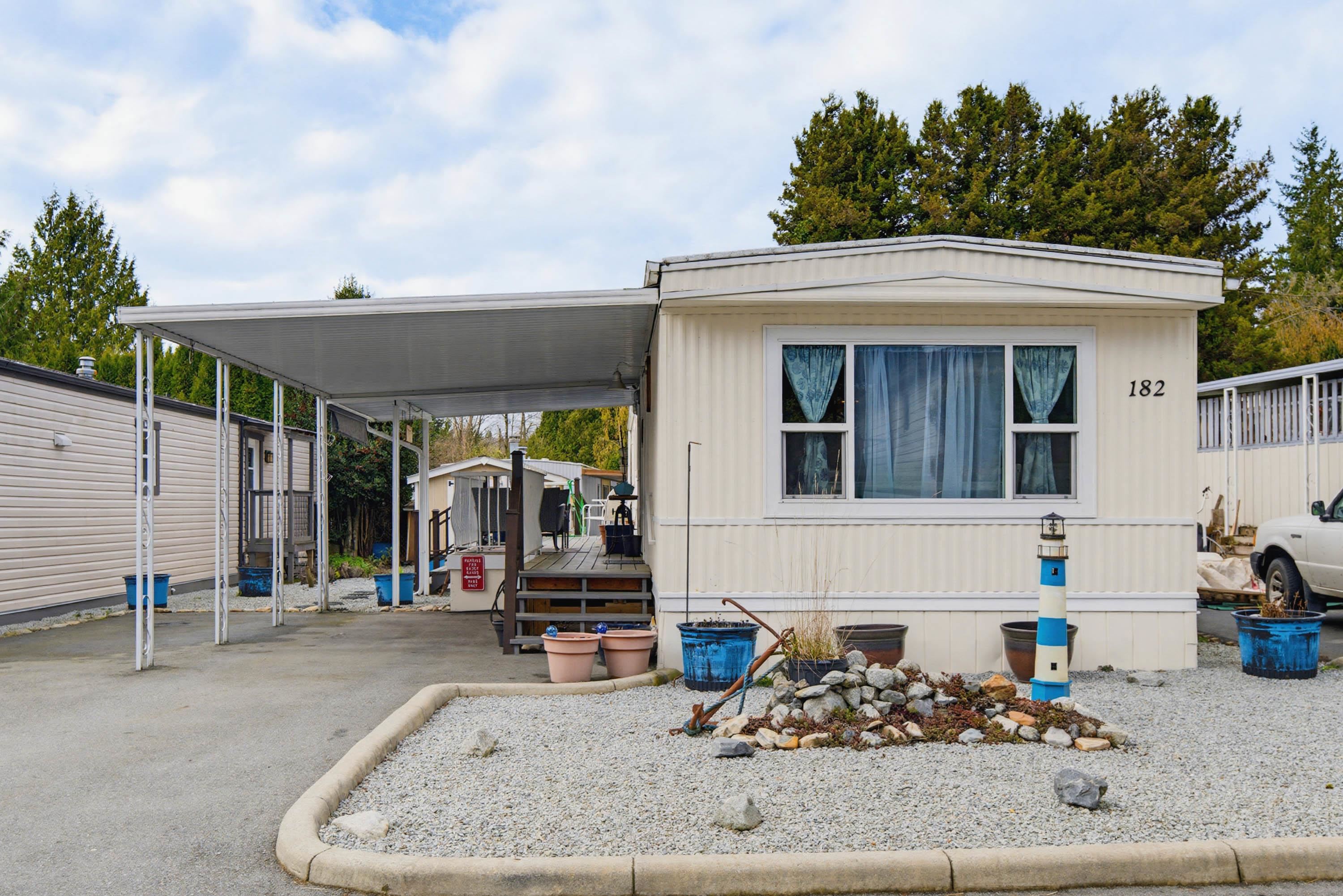 Cream manufactured home with a wide carport, gravel landscaping, and number 182 beside the front window.