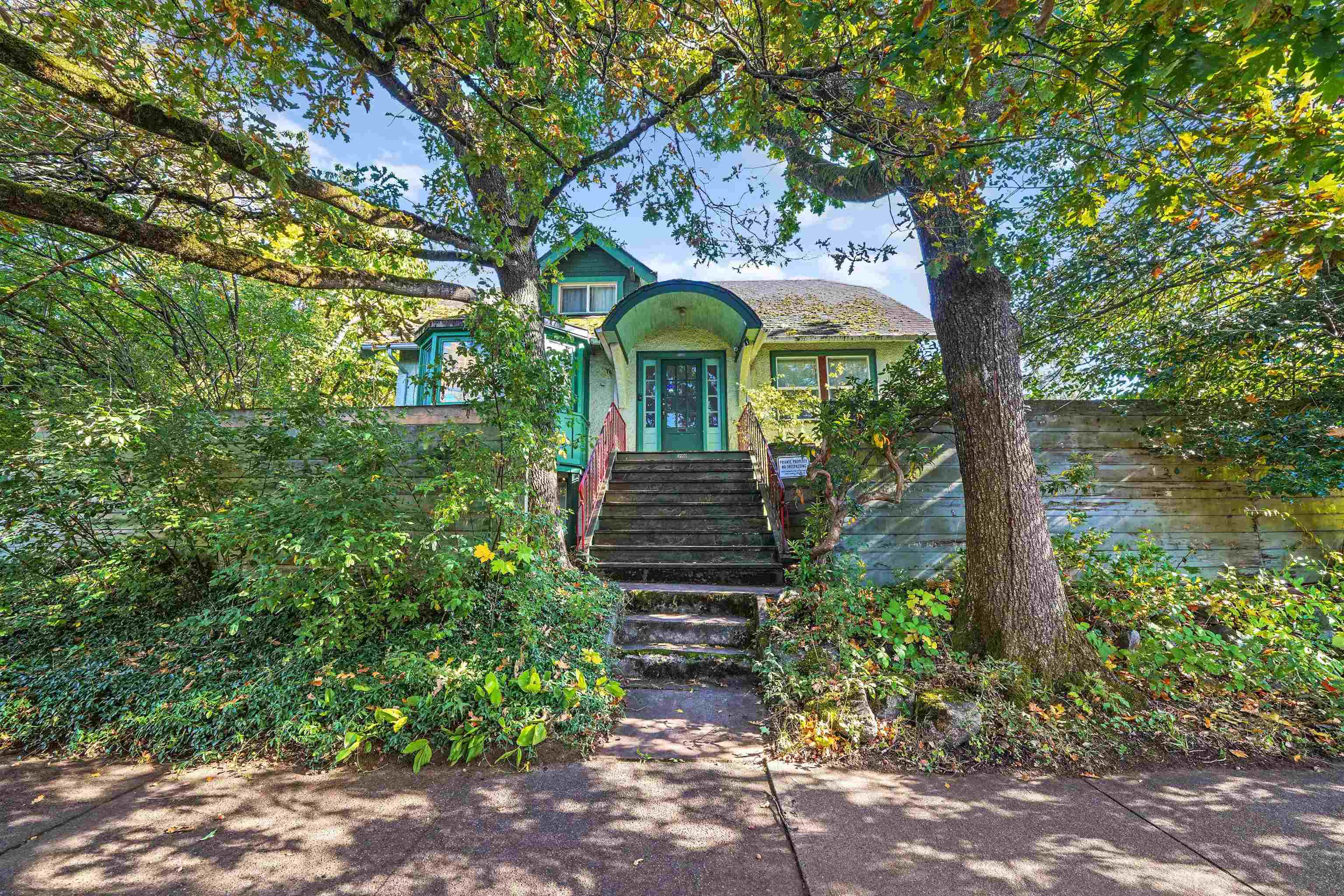 House with teal trim and an arched entry, framed by mature trees and dense greenery.