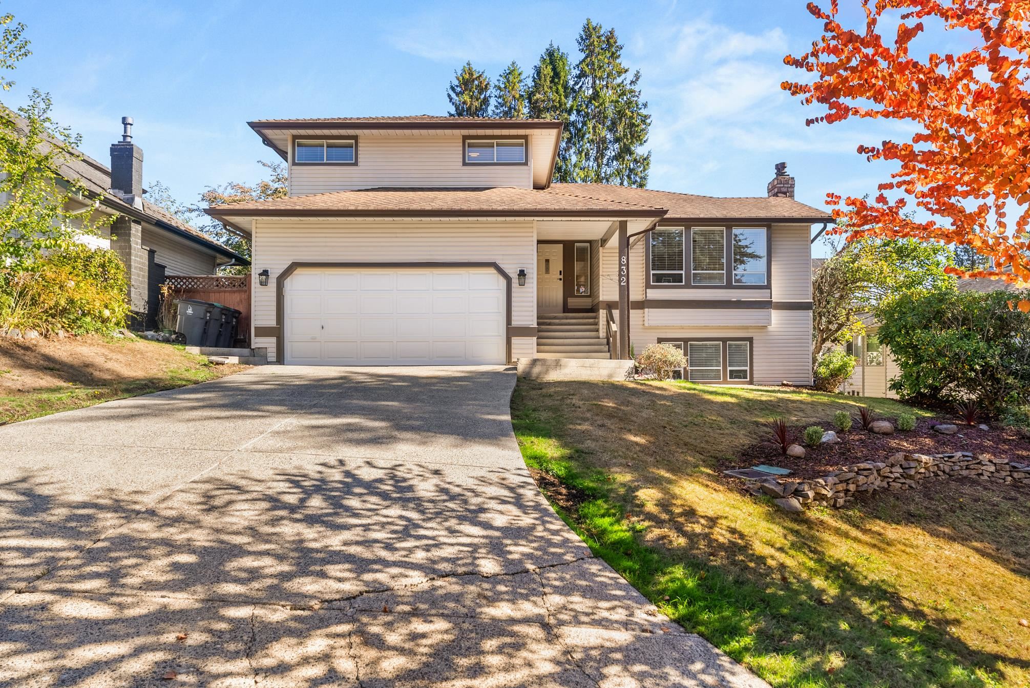 Beige split-level house with a double garage, wide driveway, and orange autumn leaves.