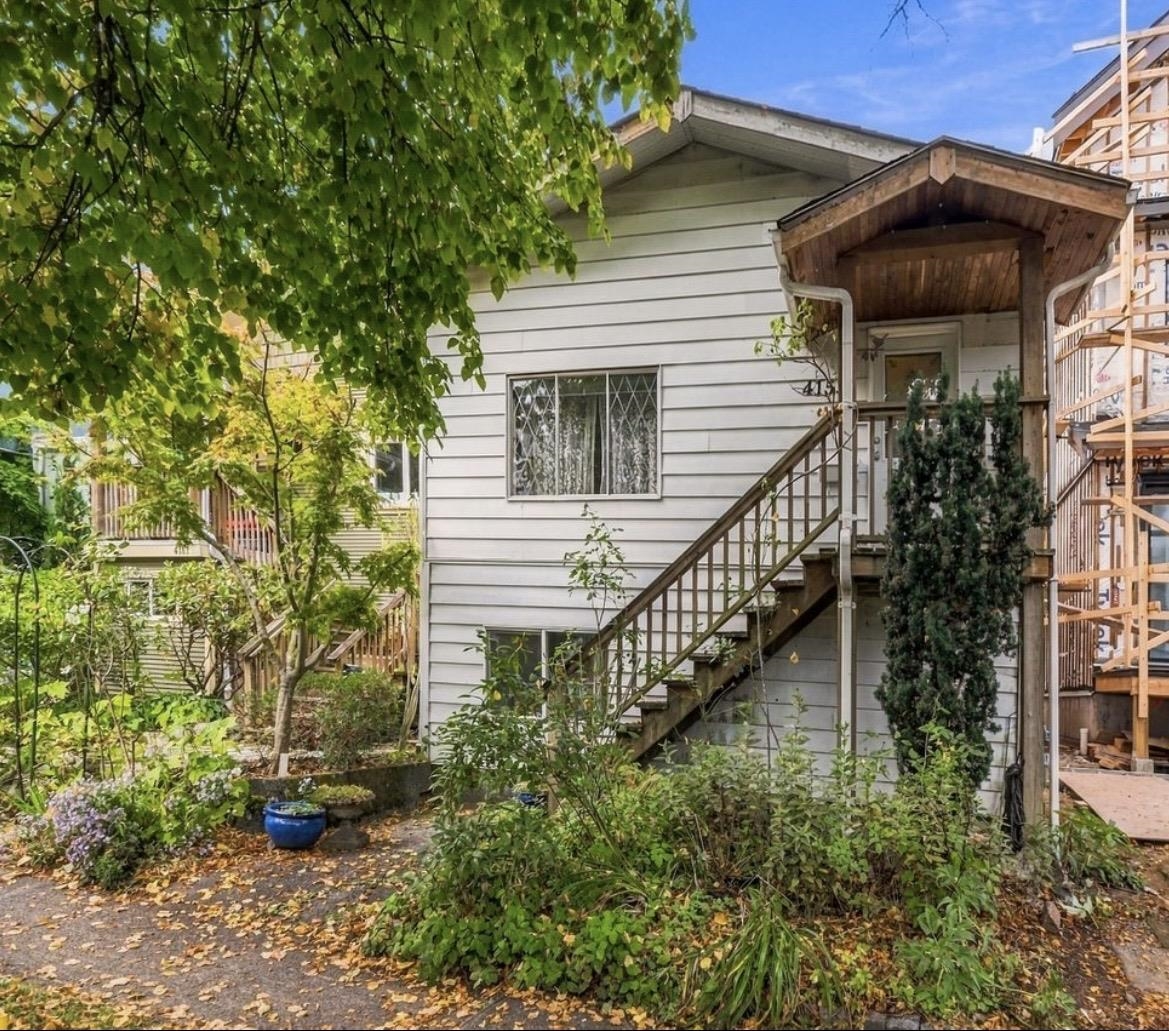 White-sided home with a covered wooden entry and exterior staircase, framed by leafy trees and garden plants.