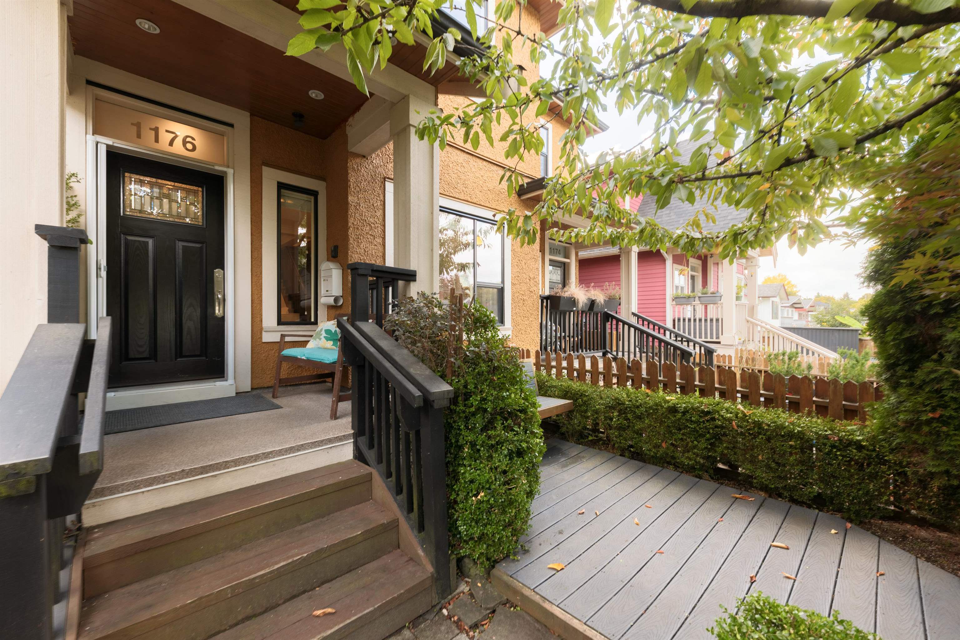 Townhouse entrance with a black door, wooden steps, and a gray deck beneath leafy trees.