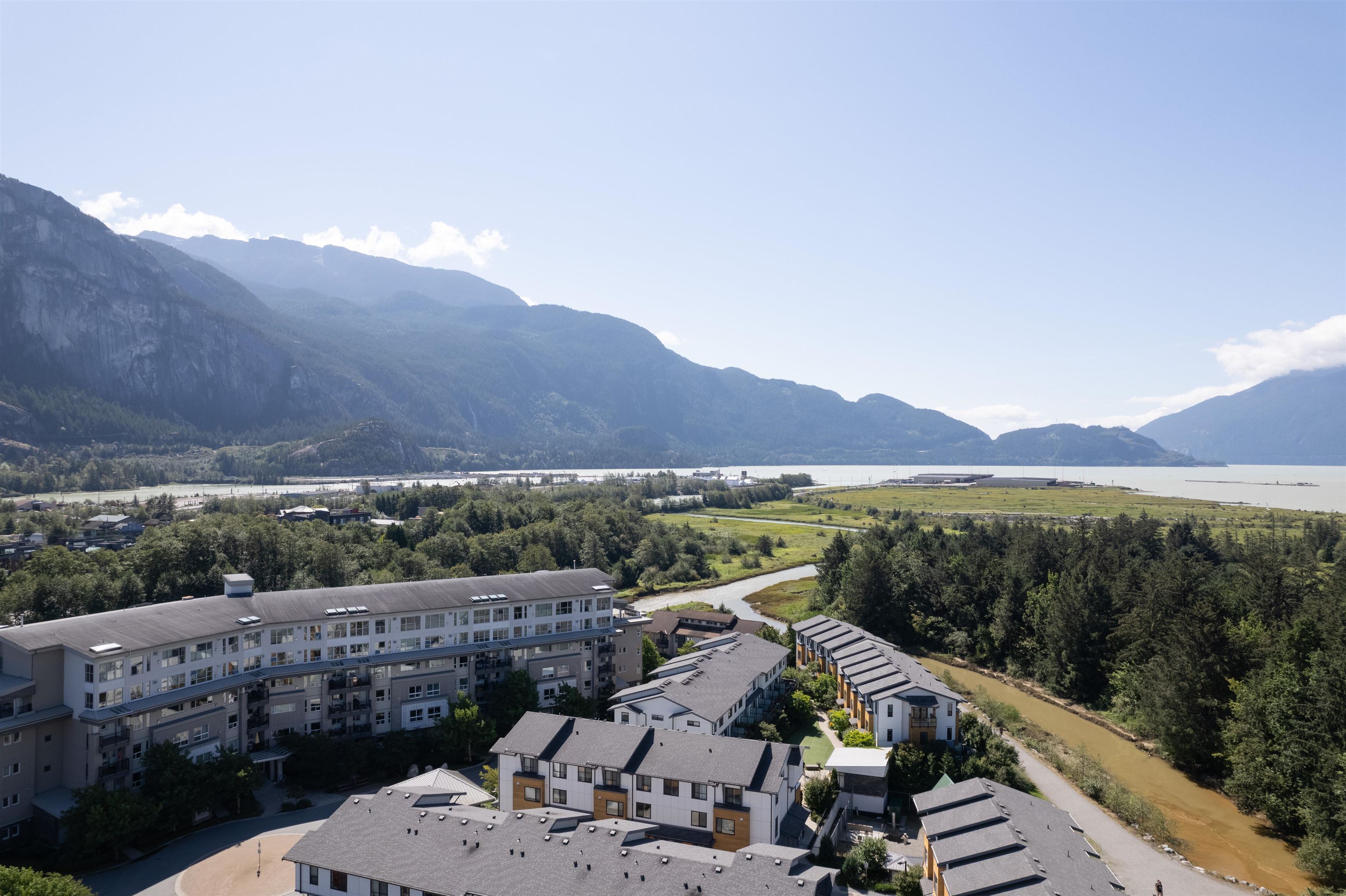 Aerial view of residences beside a river, with forested mountains and water beyond.