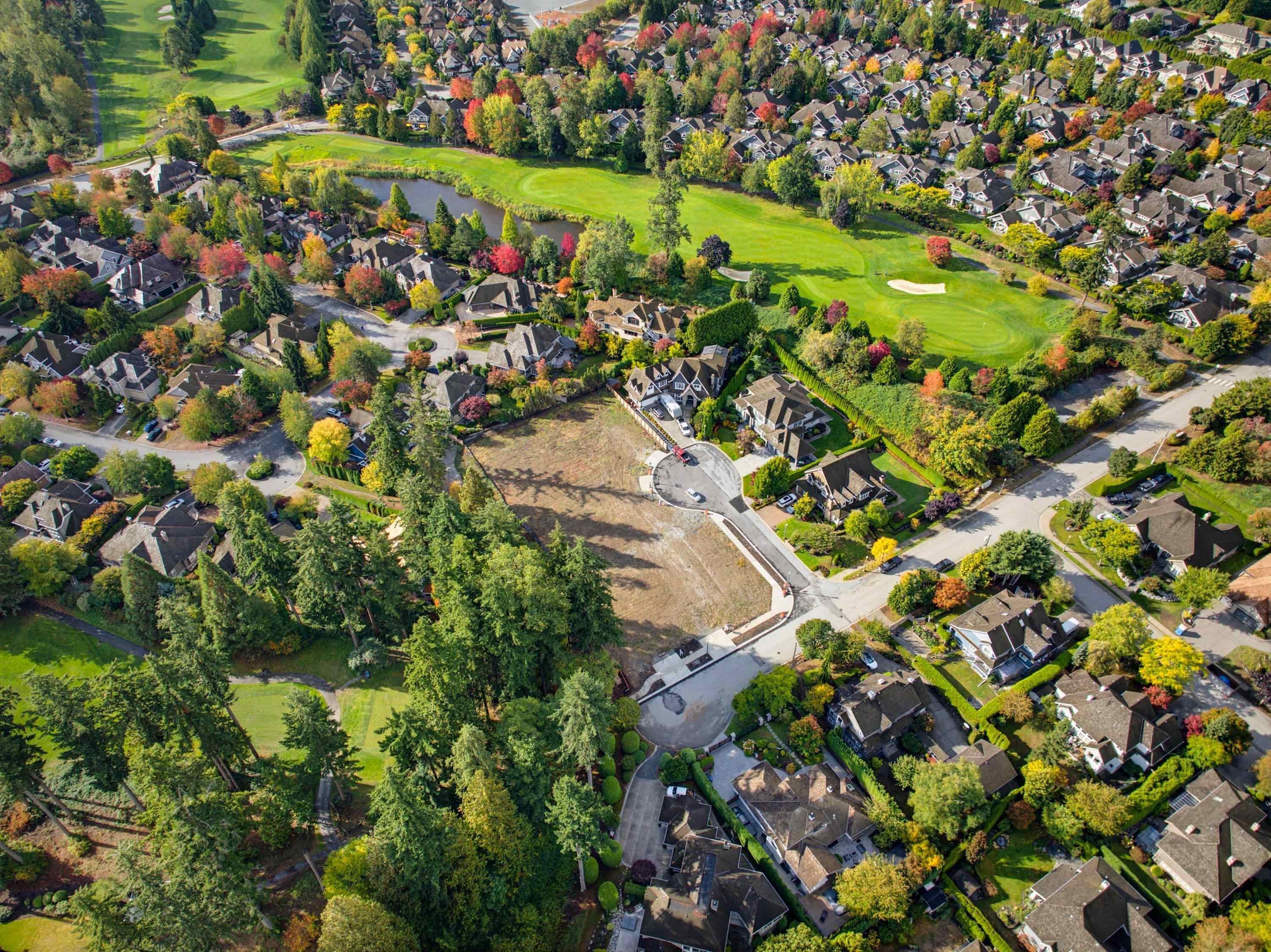 Aerial neighborhood with a golf course, pond, and cleared lot among autumn trees.