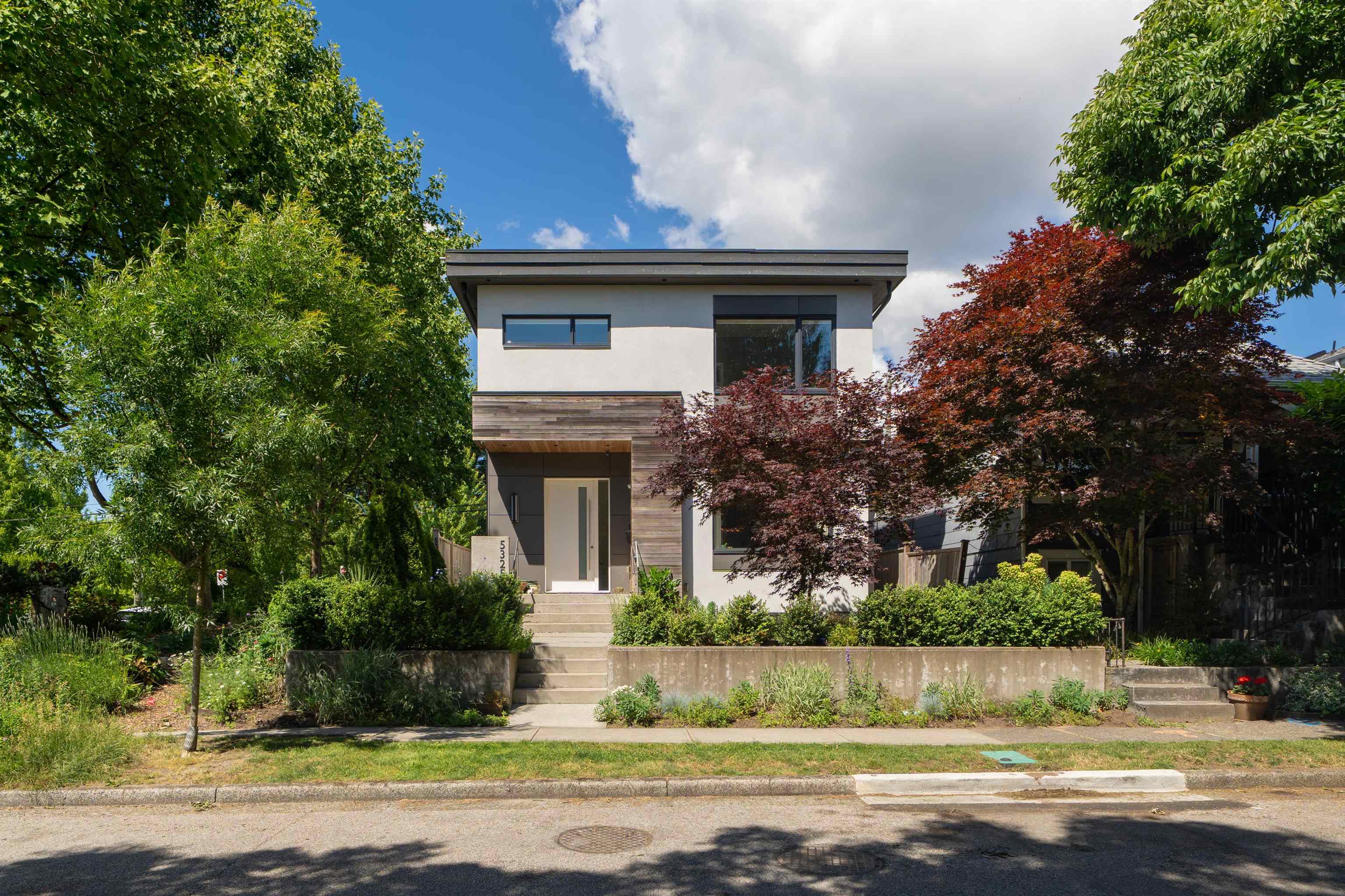 Modern two-story house with a wood-clad entry, front steps, and mature landscaped garden.