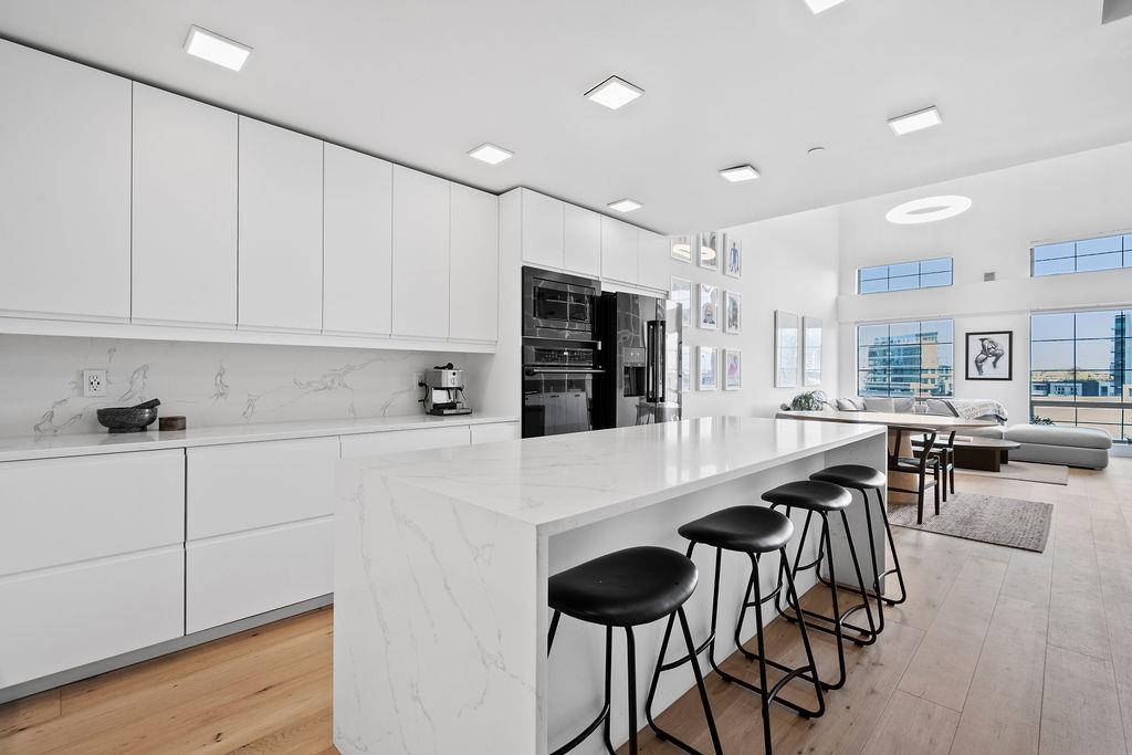 Modern white kitchen with a long island and black stools opening onto a windowed living area.