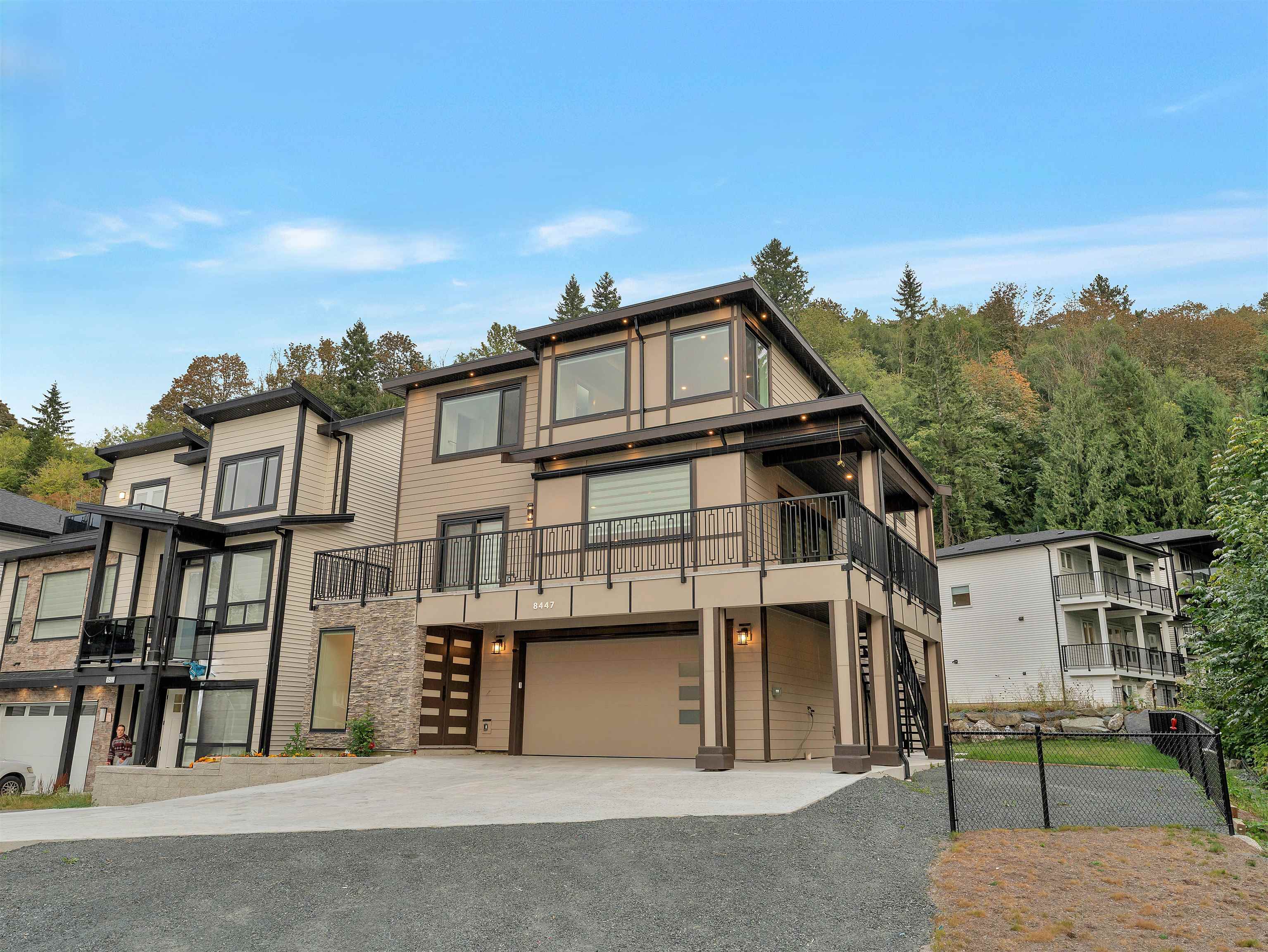 Modern multi-level house with a broad balcony and attached garage, backed by a wooded hillside.
