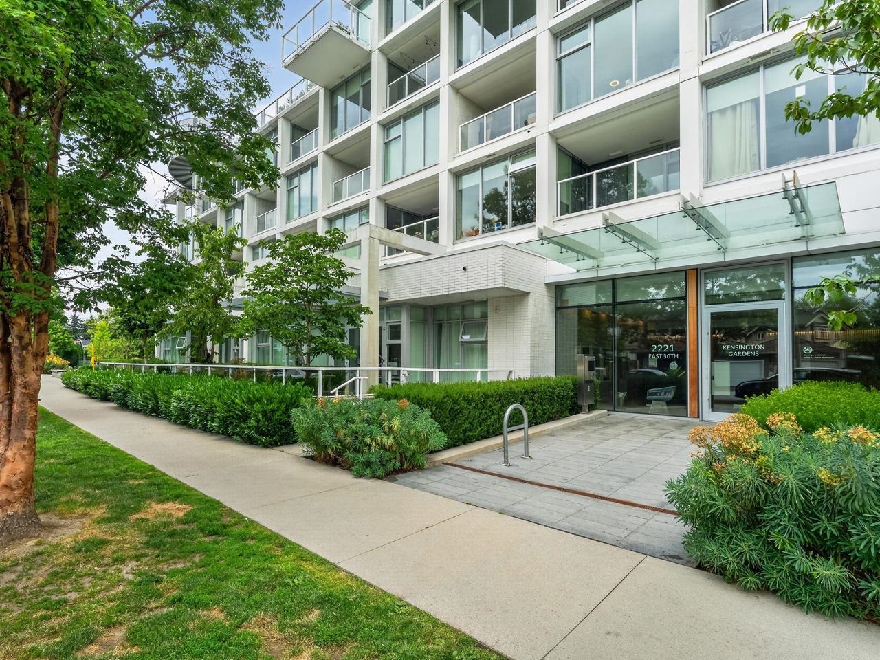 Kensington Gardens condominium entrance with glass canopy, landscaped shrubs, and sidewalk.
