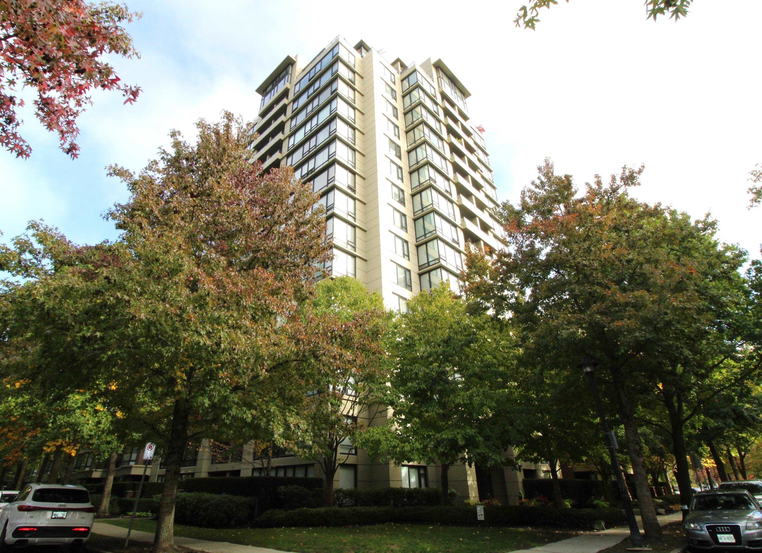 Beige condominium tower with balconies rising above autumn trees along a Richmond street.