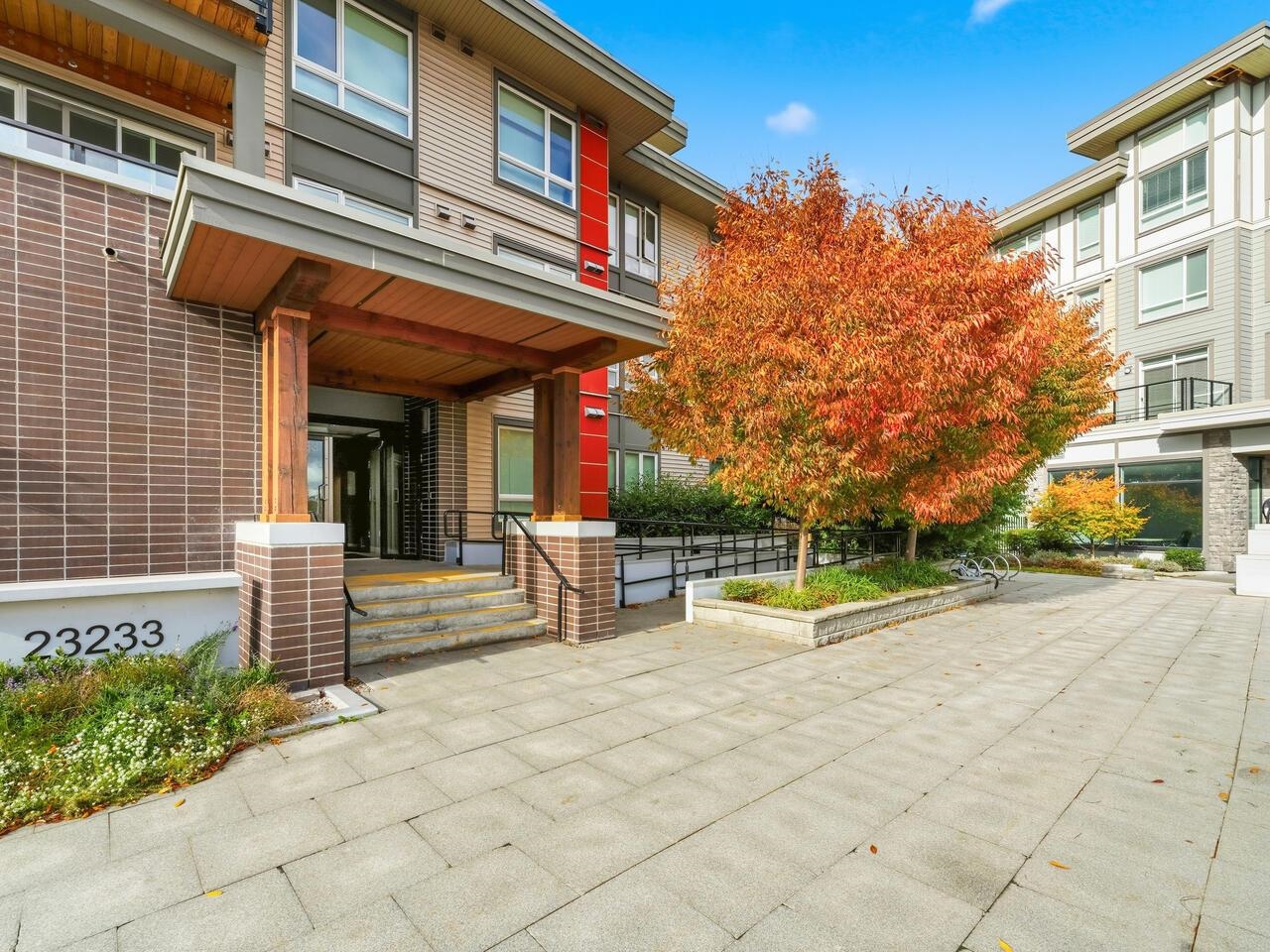 Modern residential building entrance with wood canopy, paved courtyard, and orange autumn trees.