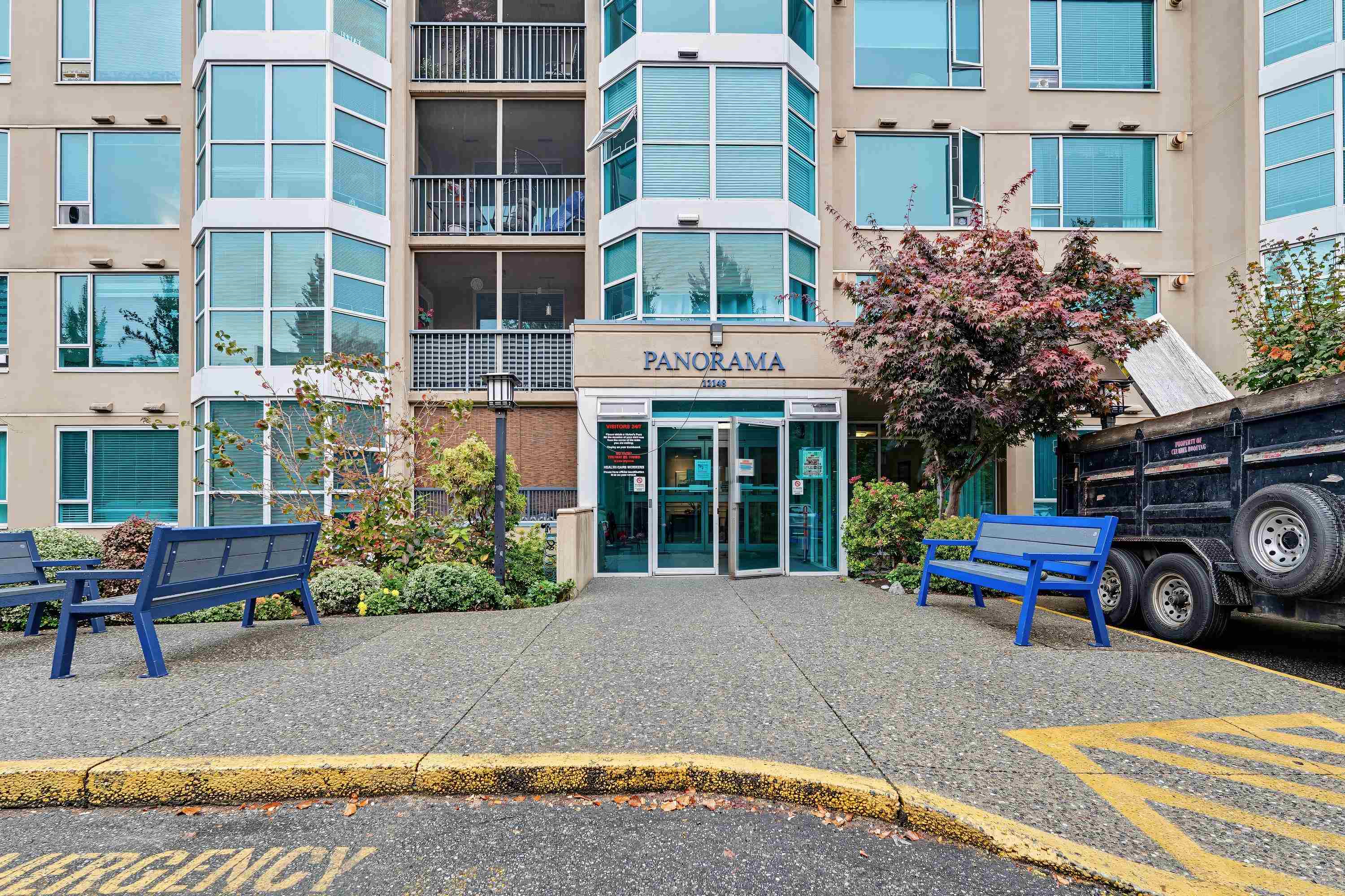 Panorama condominium entrance with projecting bay windows, landscaped shrubs, and blue benches.
