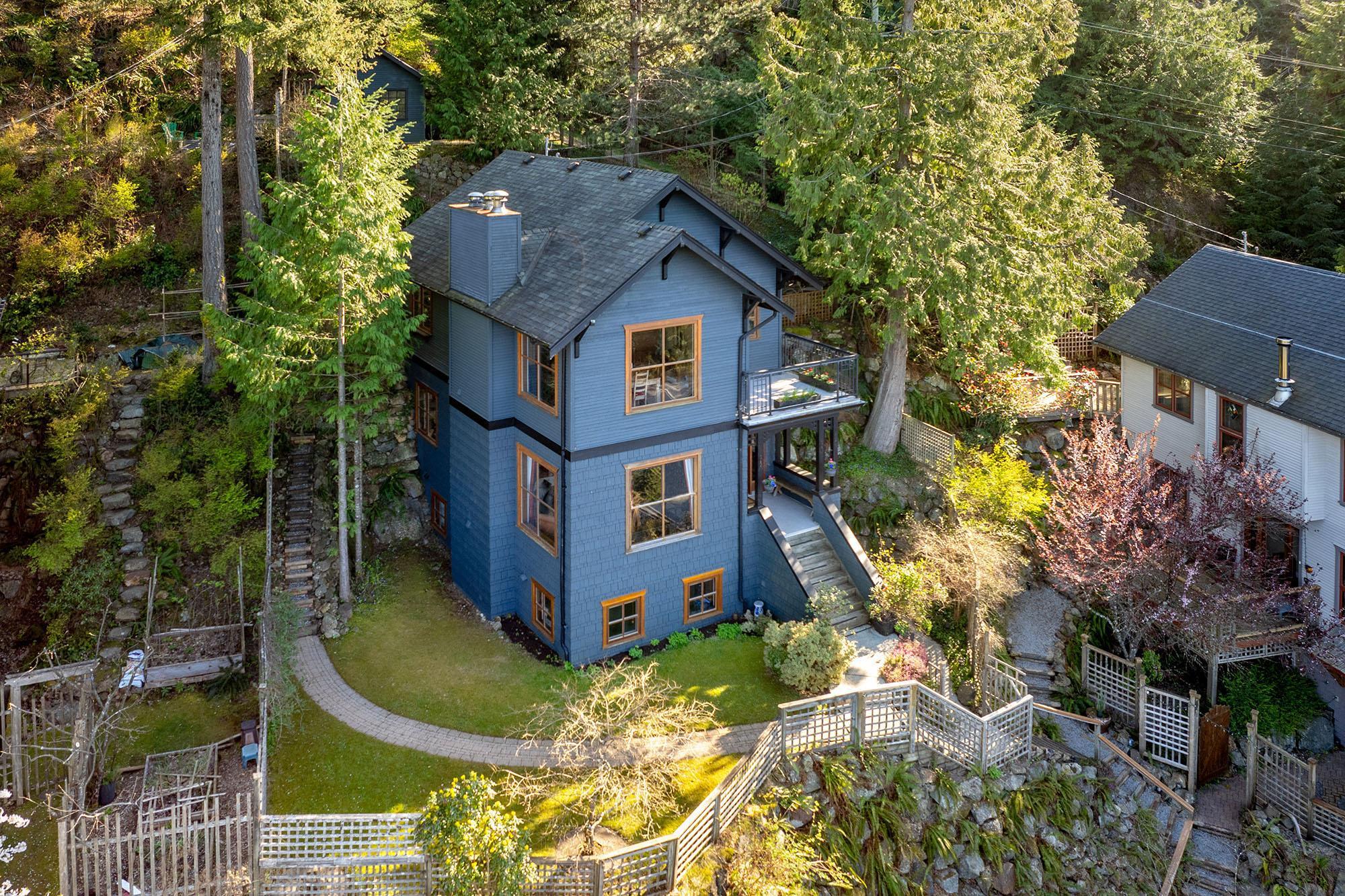 Blue multi-level home with wood-trimmed windows, a balcony, and a landscaped forested hillside lot.