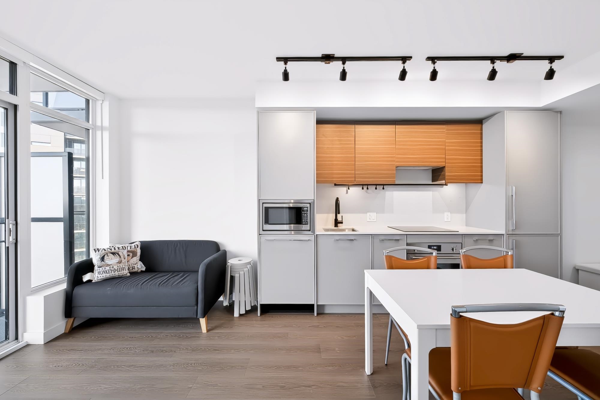 Modern open-plan kitchen and living area with wood cabinets, a white dining table, and sofa beside large windows.