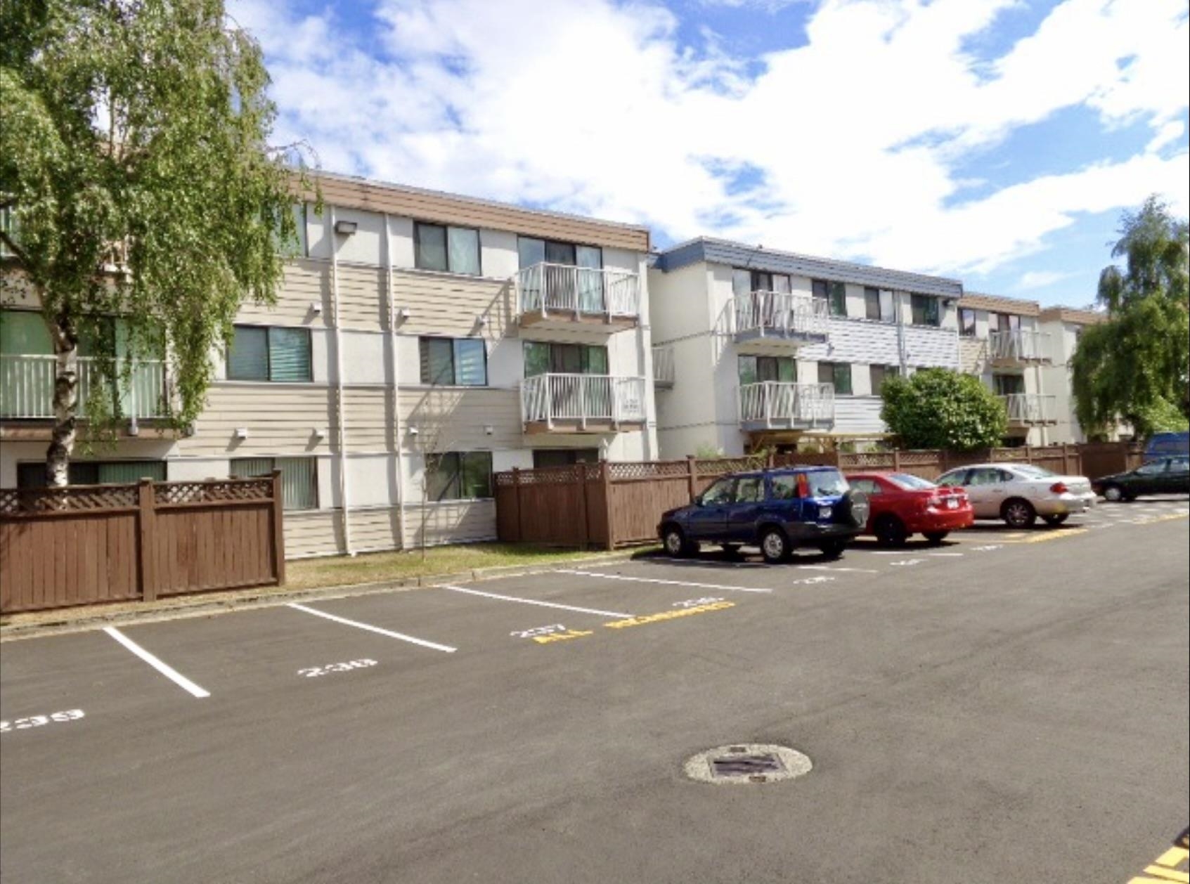 Low-rise apartment building with private balconies and fenced ground-floor patios along a parking lot.