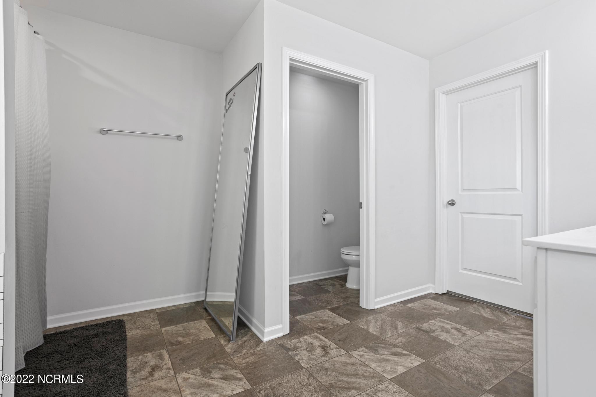 Bathroom with a tiled floor, shower curtain, leaning full-length mirror, and toilet visible through an open doorway.