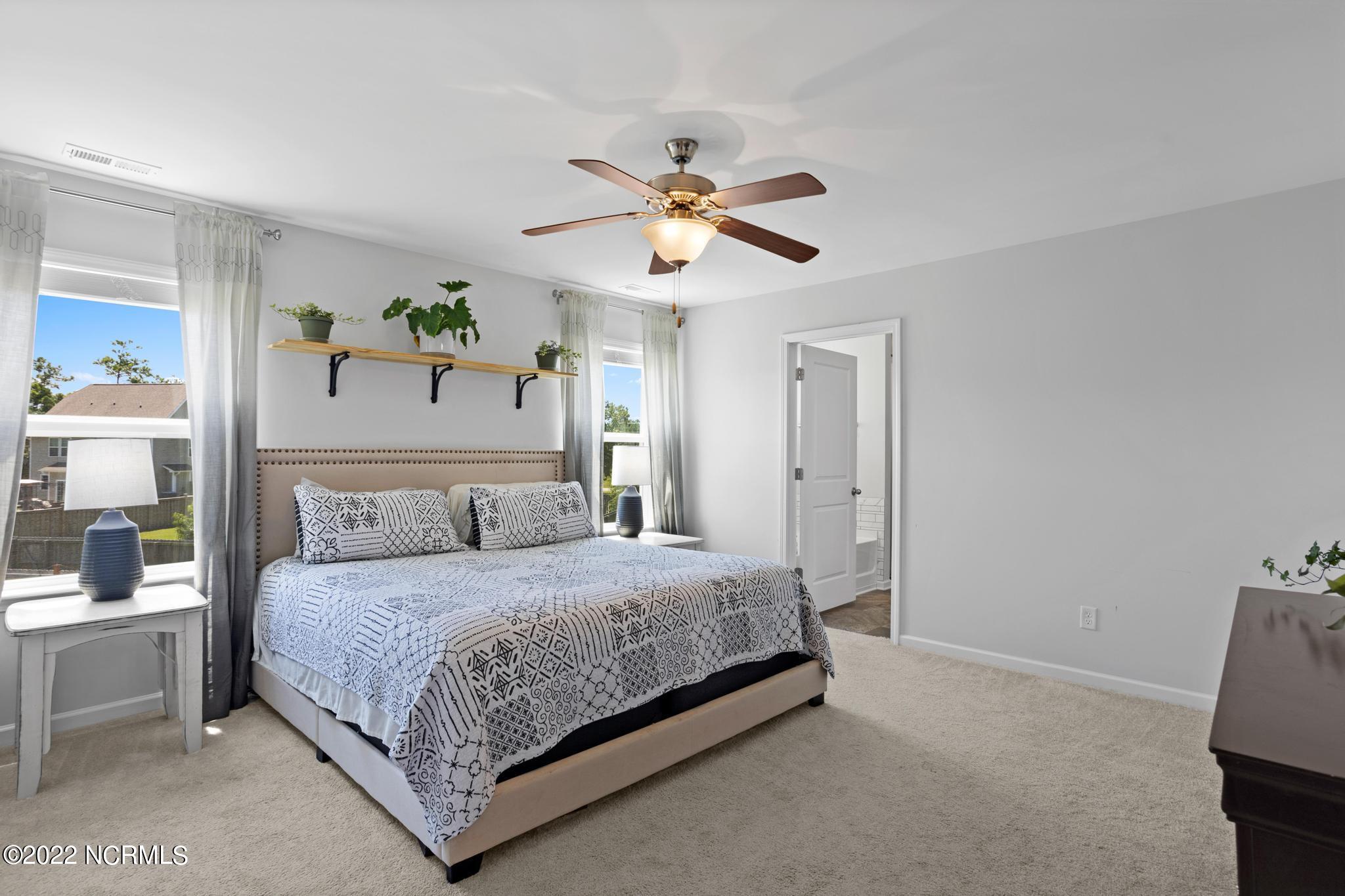 Bedroom with patterned bedding, a ceiling fan, two windows, and plants on a wall shelf.