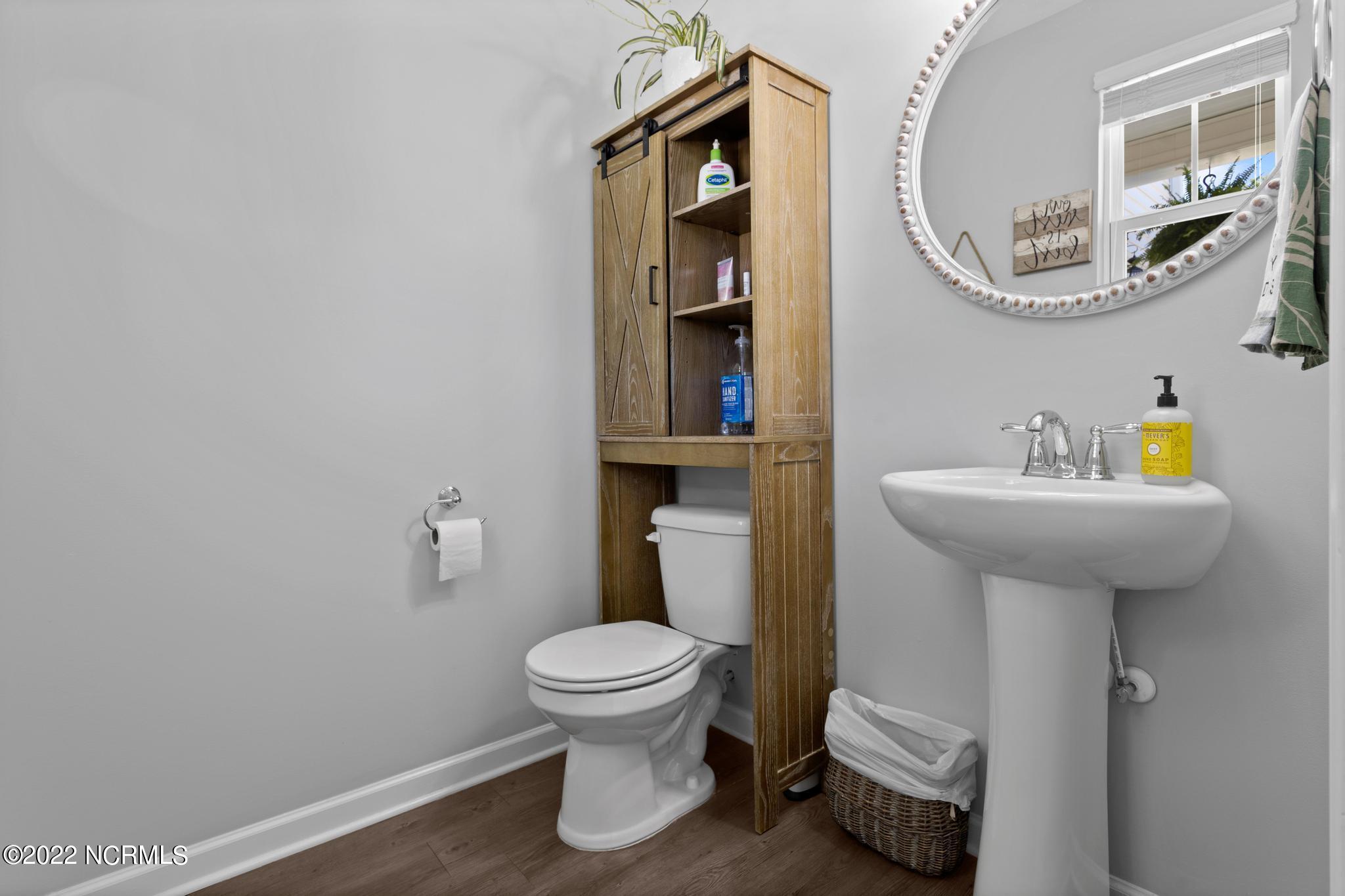 Bathroom with a white pedestal sink, toilet, rustic wood storage cabinet, and large round mirror.