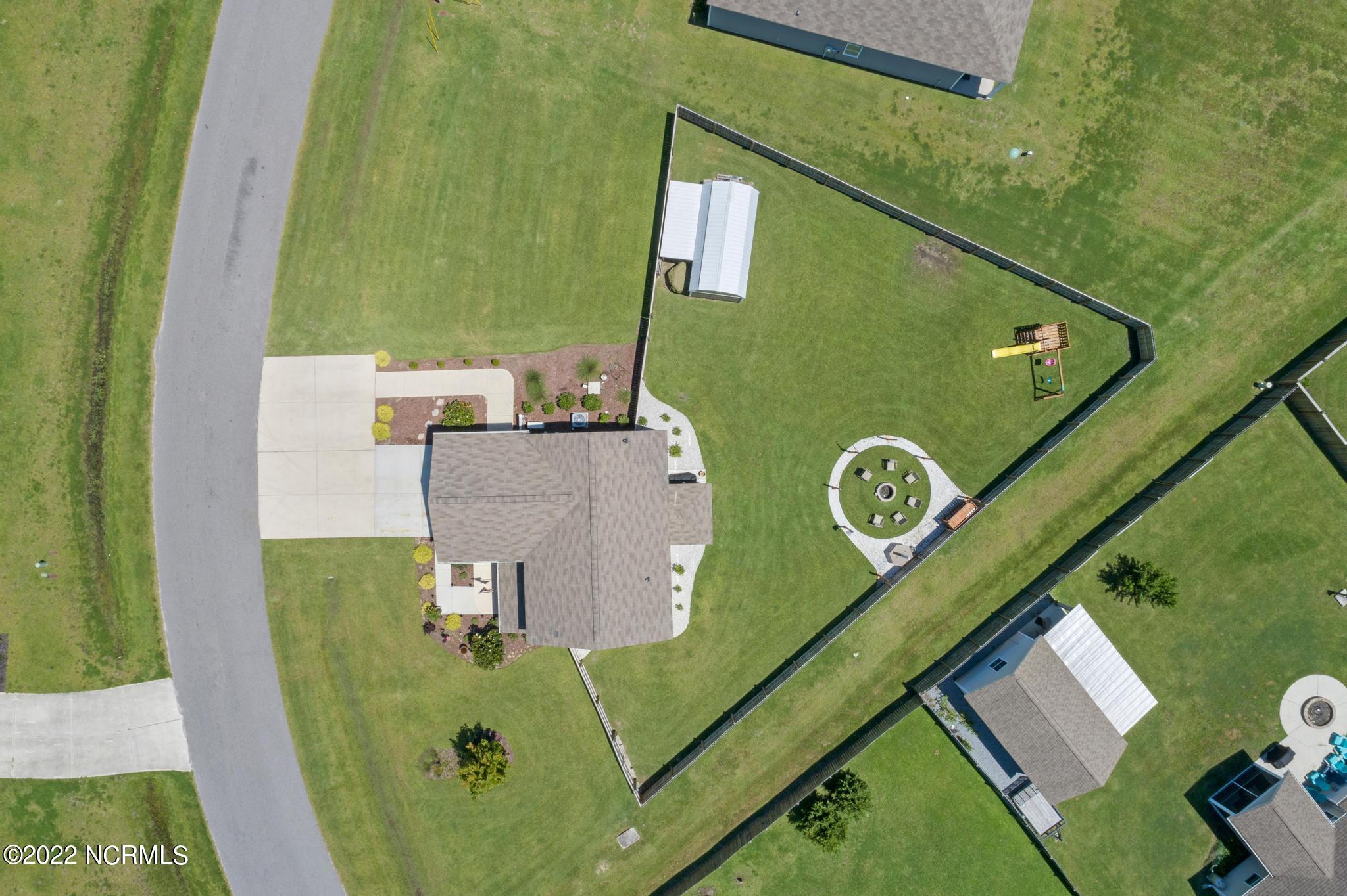 Aerial view of a home with a fenced lawn, circular fire pit area, playset, and outbuilding.