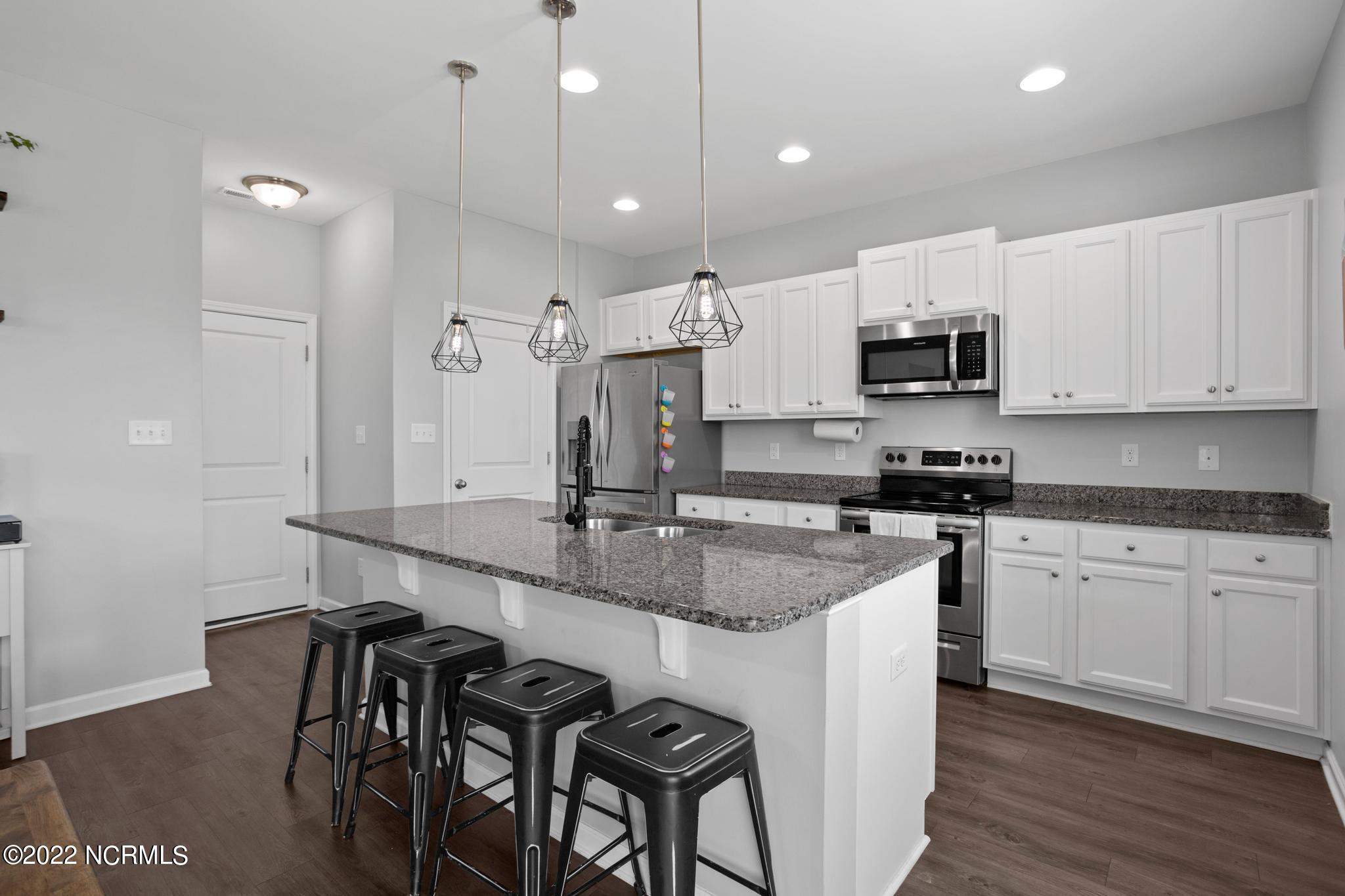 Kitchen with a granite island, four black stools, white cabinets, and stainless steel appliances.