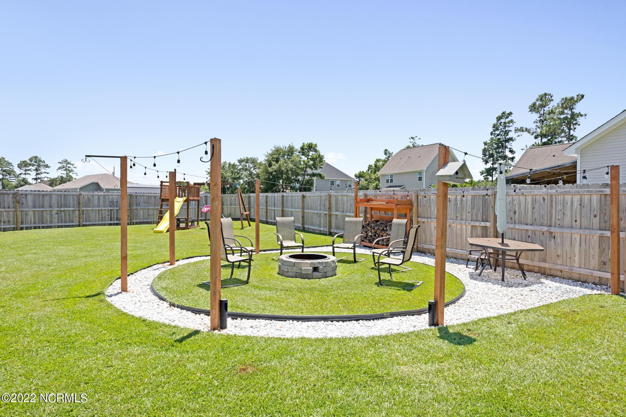 Fenced backyard with a circular fire pit seating area, string lights, and a children’s playset.