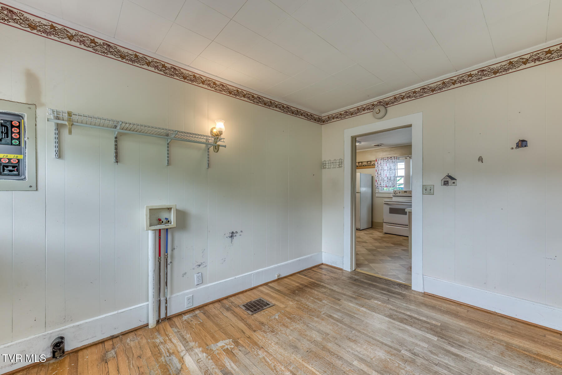 Utility room with washer hookups, a wire shelf, and worn hardwood flooring beside the kitchen doorway.