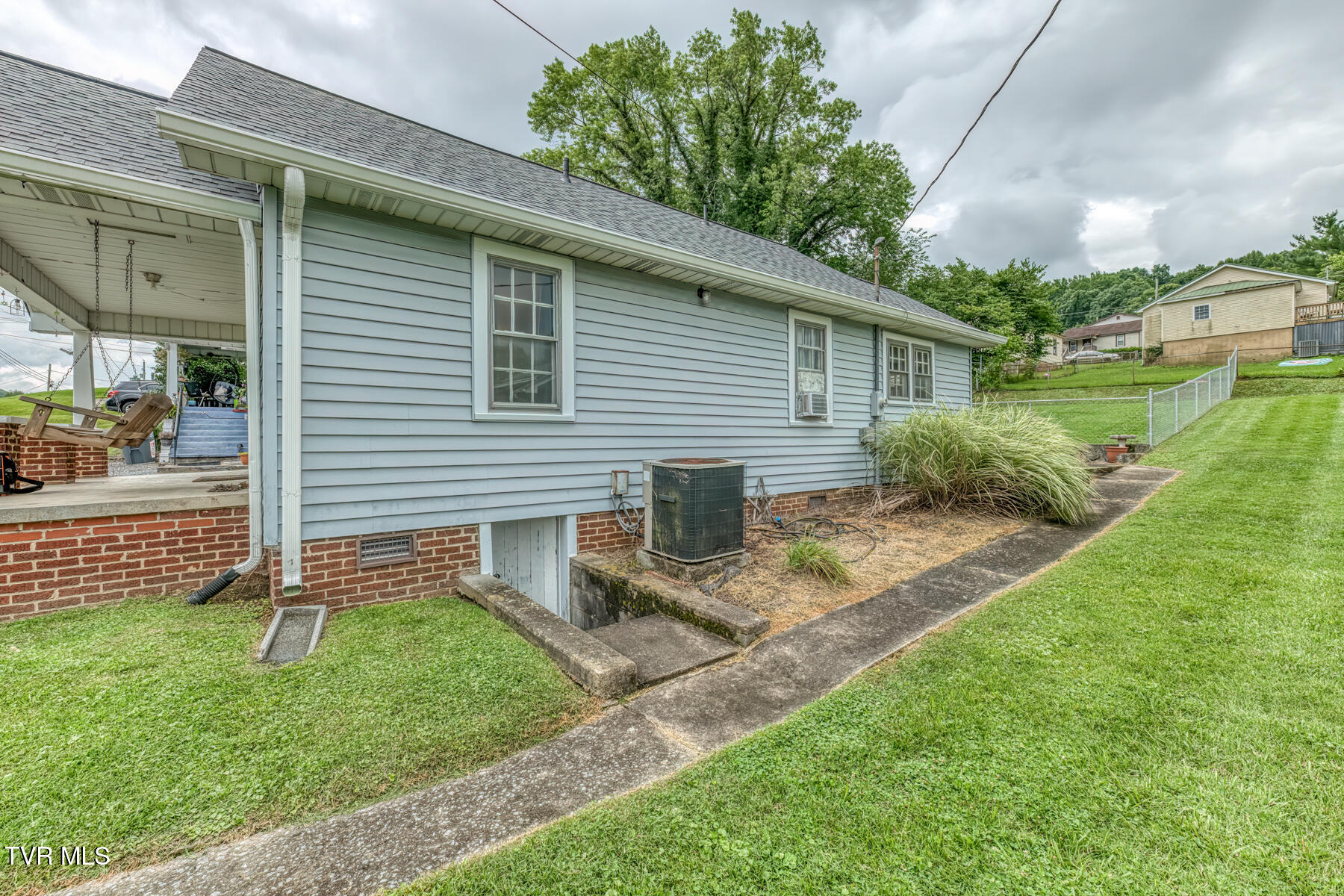 Side of a gray-sided home with covered carport, concrete walkway, lawn, and air-conditioning unit.
