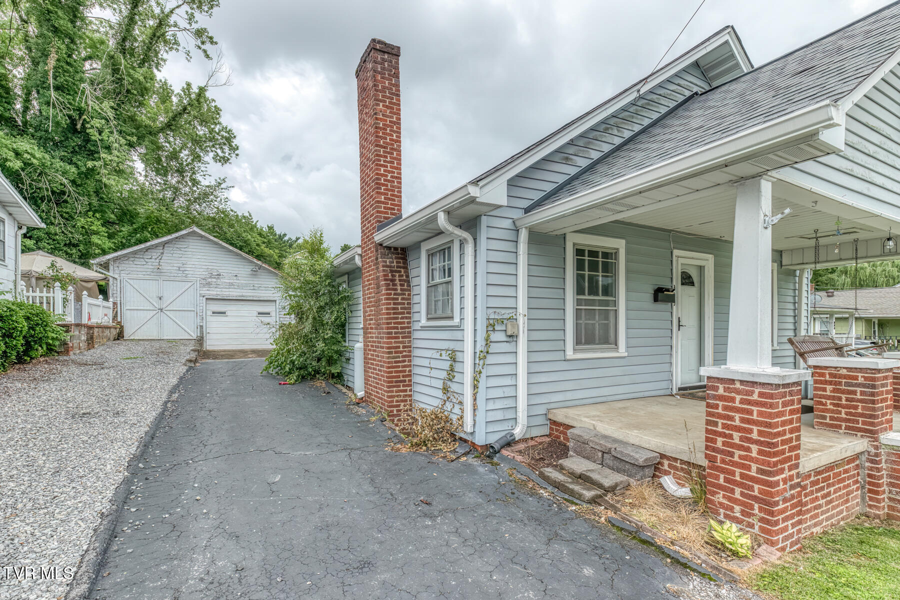 Blue-gray house with a covered porch, brick chimney, and driveway beside a detached garage.