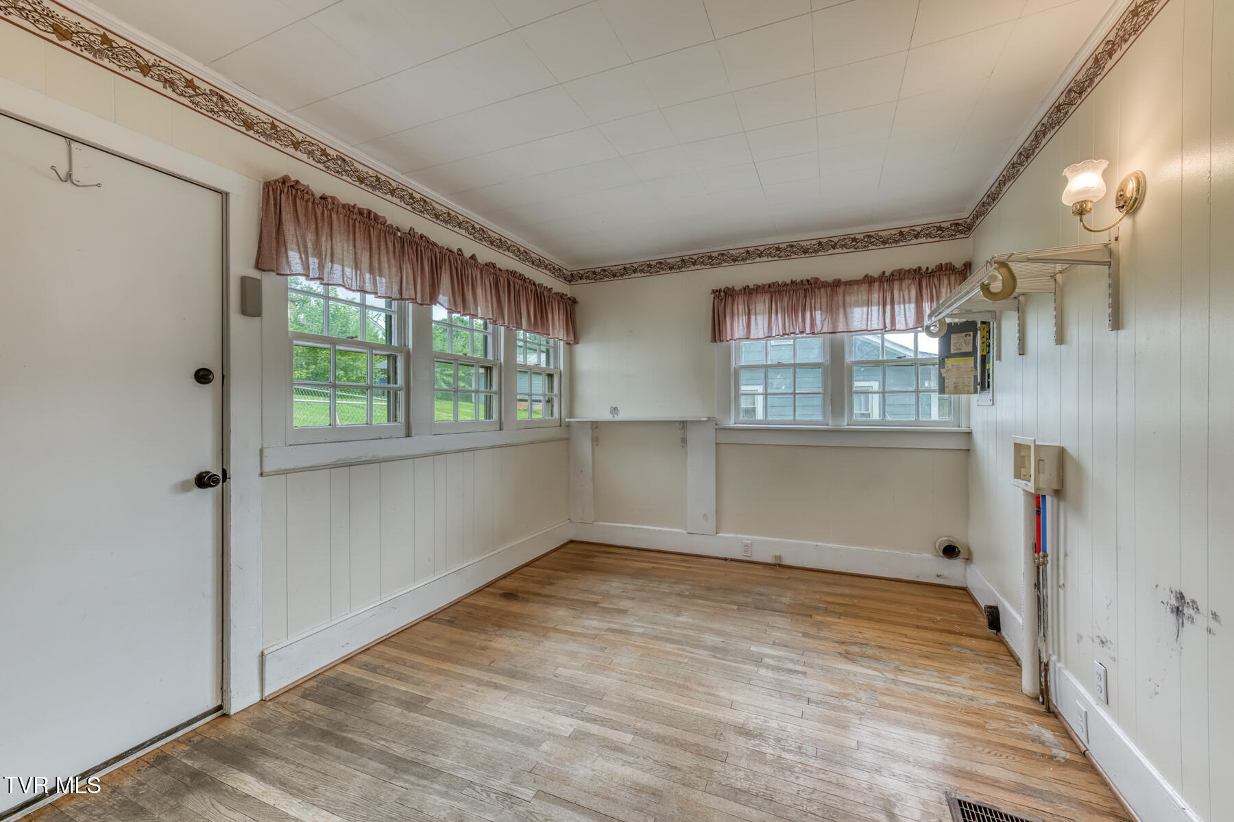 Vacant room with wood flooring, paneled walls, multiple windows, and utility hookups.