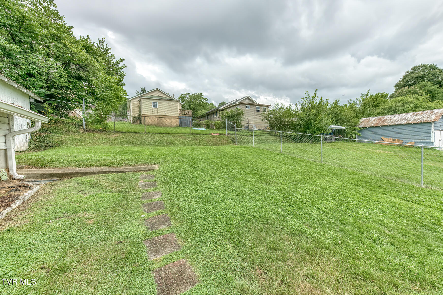 Grassy backyard with a chain-link fence, stepping stones, and mature trees.