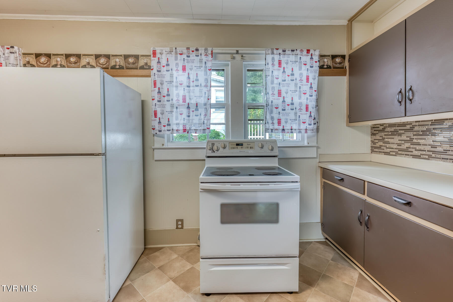 Kitchen with a white refrigerator and range, brown cabinets, and patterned curtains over the windows.