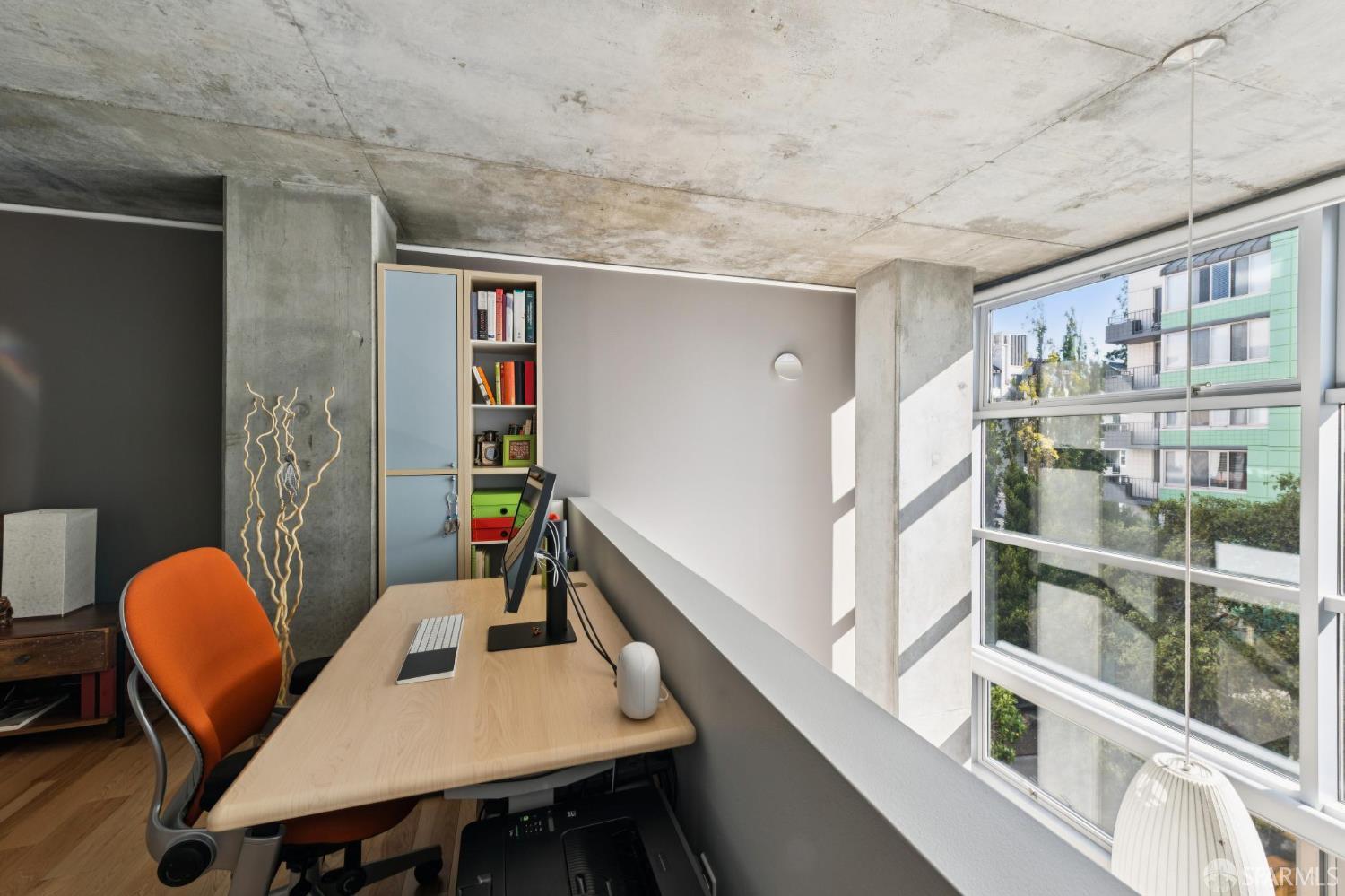 Furnished home office with an orange chair, built-in shelves, exposed concrete, and large windows.