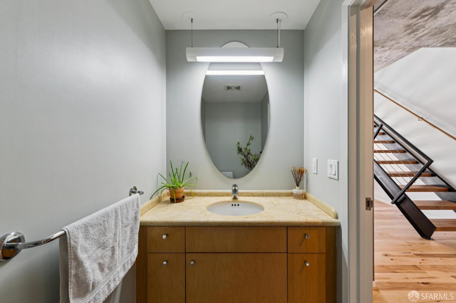 Bathroom vanity with an oval mirror, stone countertop, and wood cabinetry beside an open staircase.