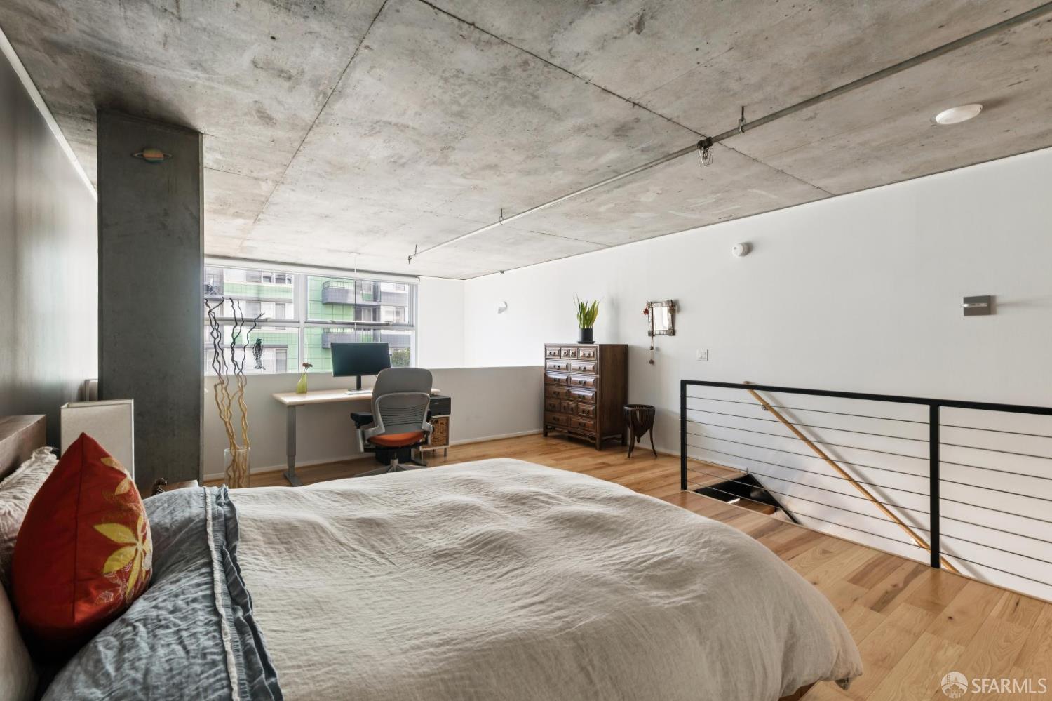 Loft bedroom with a large bed, exposed concrete ceiling, wood floors, and a desk by the windows.