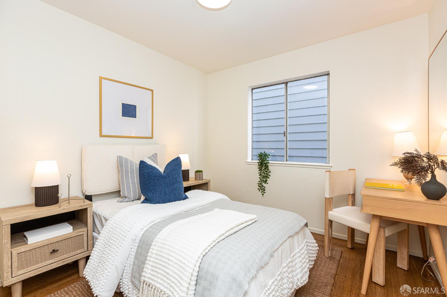 Staged bedroom with a white bed, blue accent pillow, window, and hardwood floors.