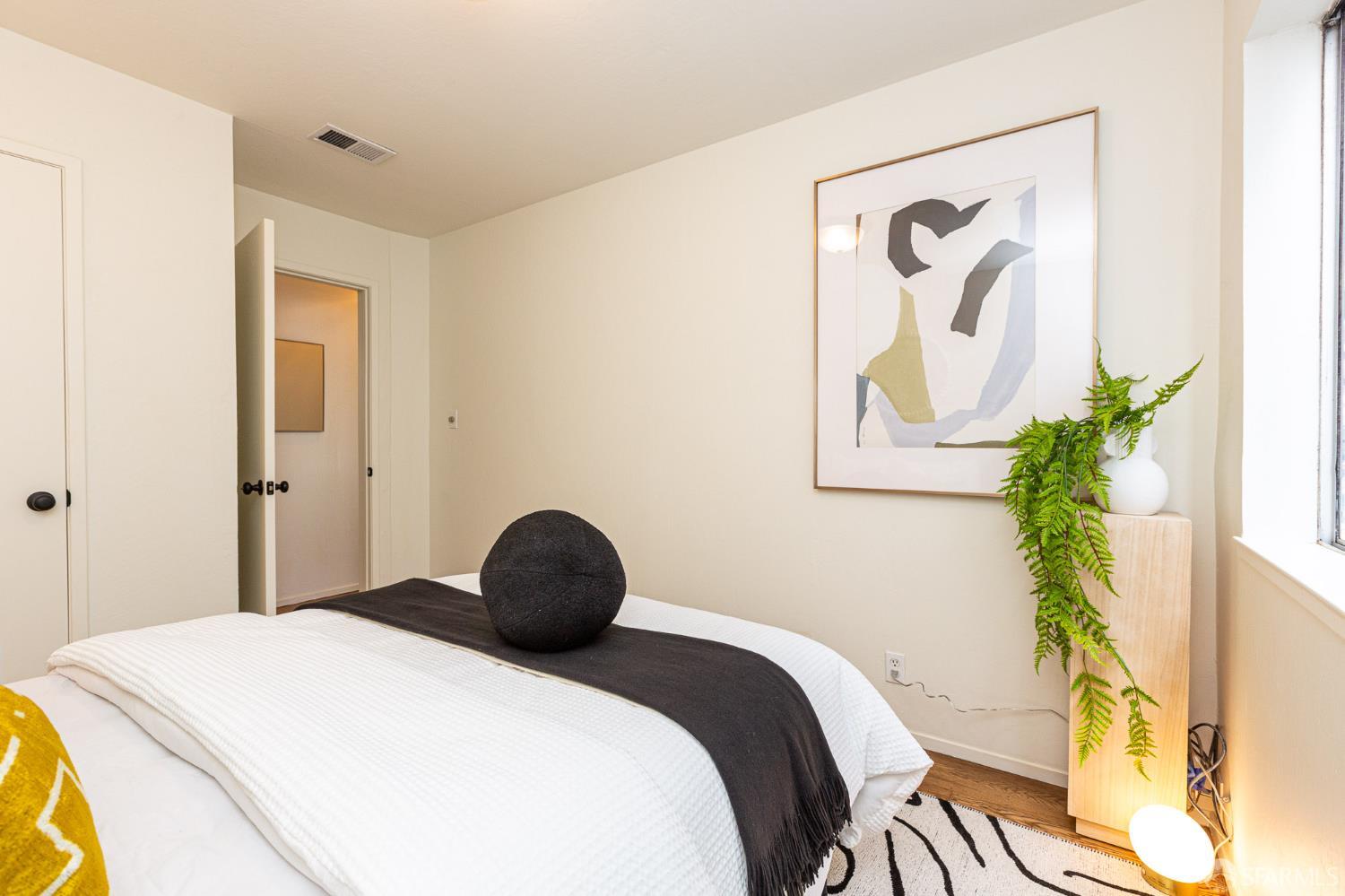 Staged bedroom with a white bed, black throw, abstract artwork, and fern beside a window.