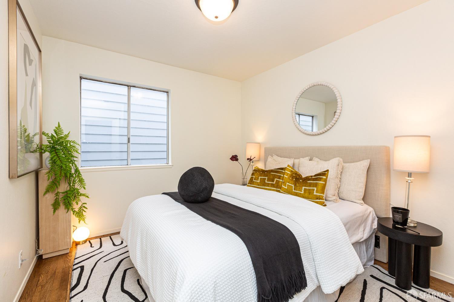 Staged bedroom with white bedding, mustard pillows, a round mirror, and a black-and-white rug.
