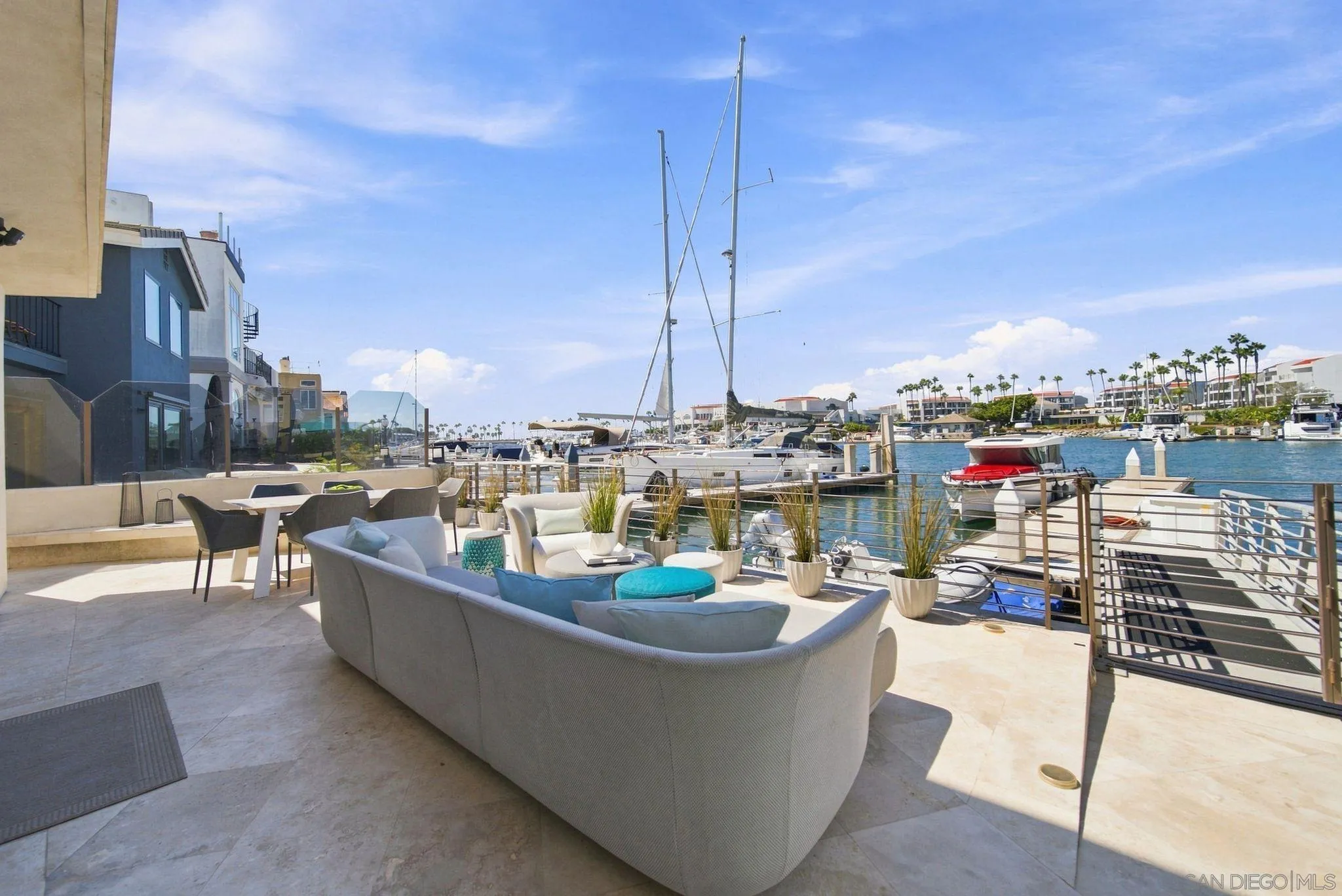 Furnished waterfront patio overlooking a marina, with lounge seating, boats, and palm trees.