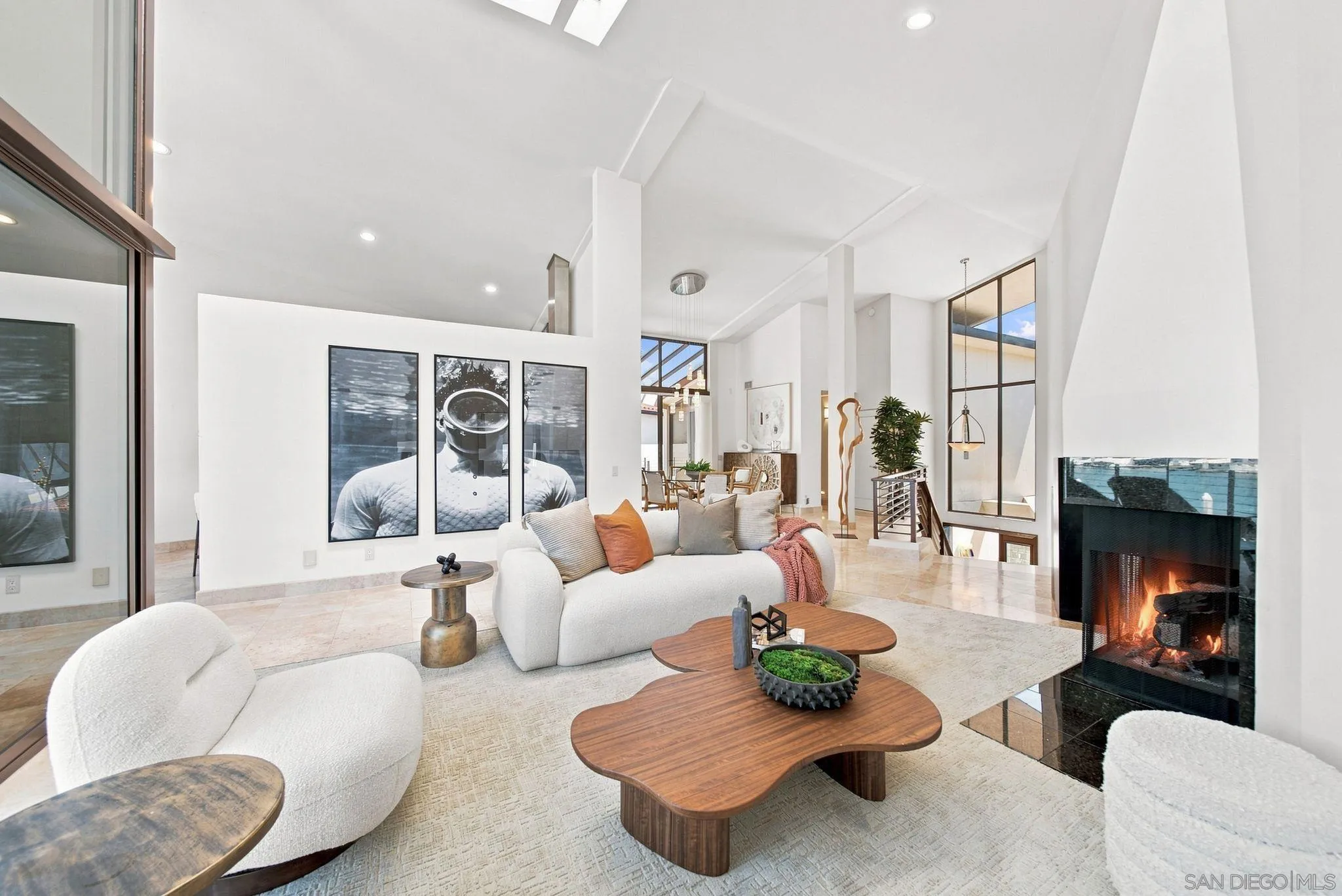 Bright living room with a vaulted ceiling, sculptural coffee tables, and a modern fireplace with water beyond.